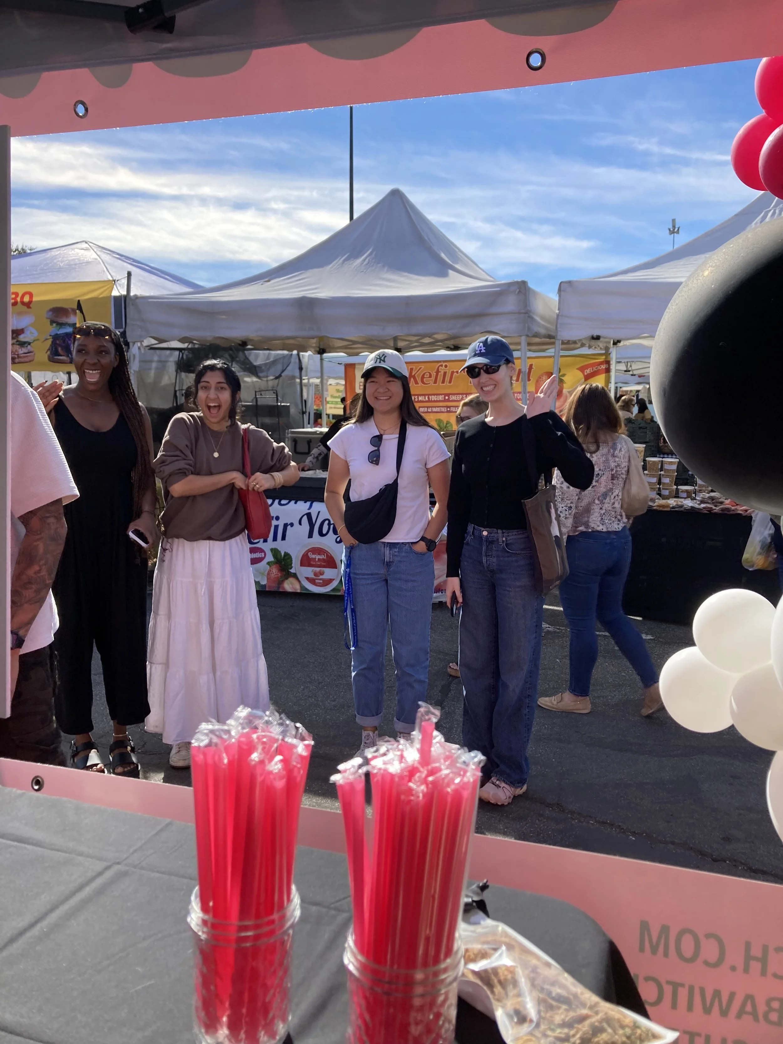 Four women smiling and standing at an outdoor market or fair, with white tents and food stalls in the background. The image is taken from inside a pink booth with pink candles and a balloon arrangement visible in the foreground.