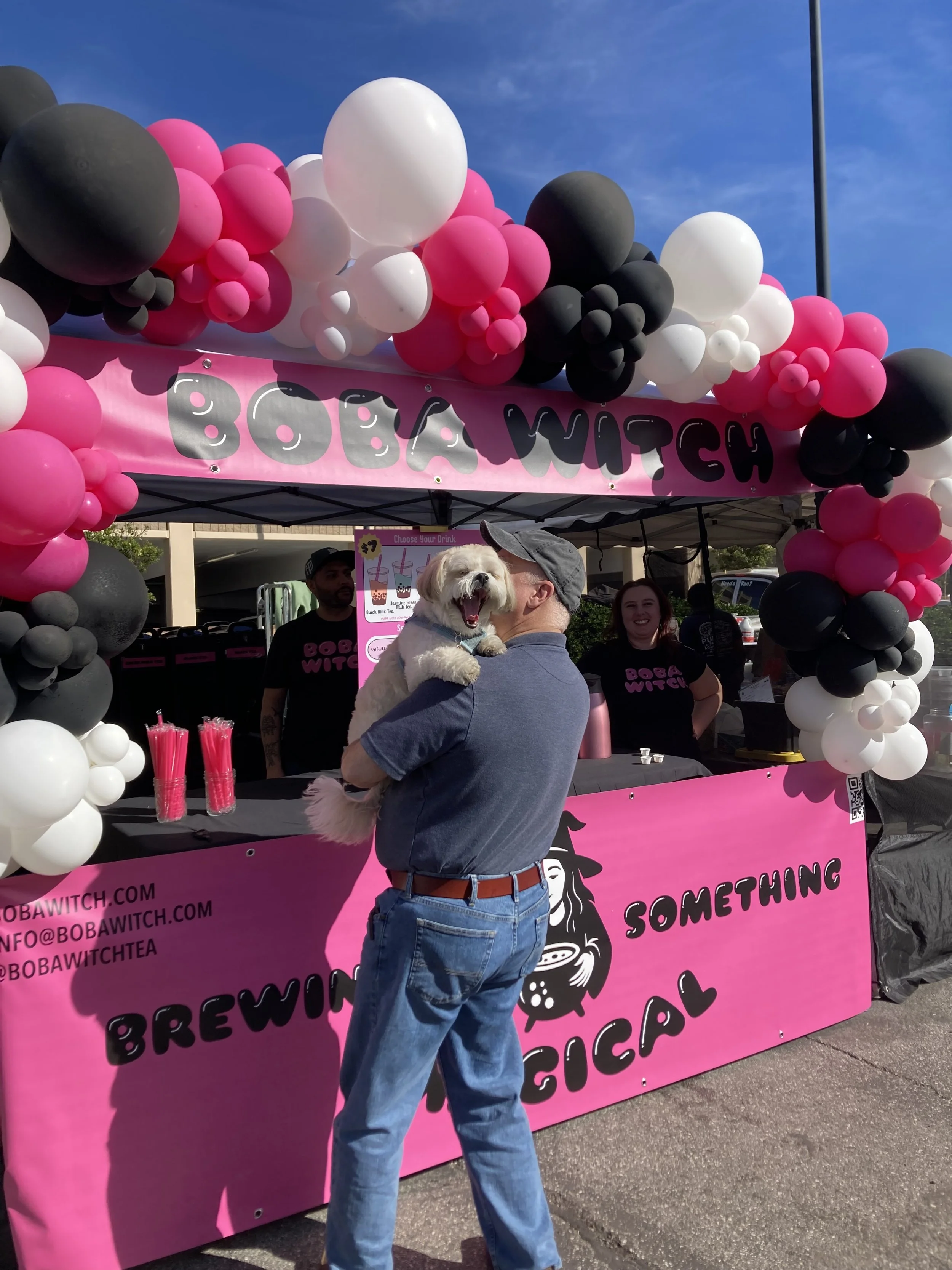 A man holding a small white dog in front of a pink and black balloon-decorated booth at an outdoor event. The booth has a pink banner with black text that reads 'BOBA WITCH' and additional information.