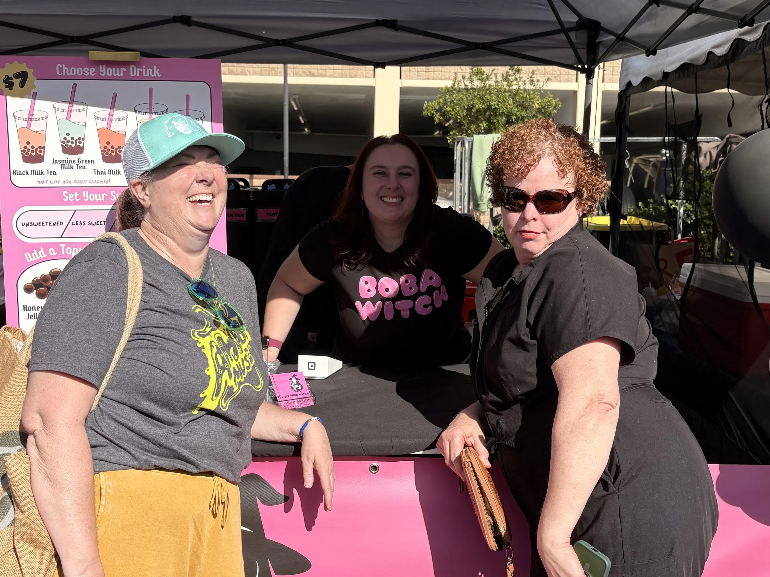 Three women at a bubble tea stand outside. The woman on the left wears a gray t-shirt and yellow pants, laughing. The woman in the middle wears a black shirt with pink letters that say "BOBA WITCH" and is smiling. The woman on the right wears a black