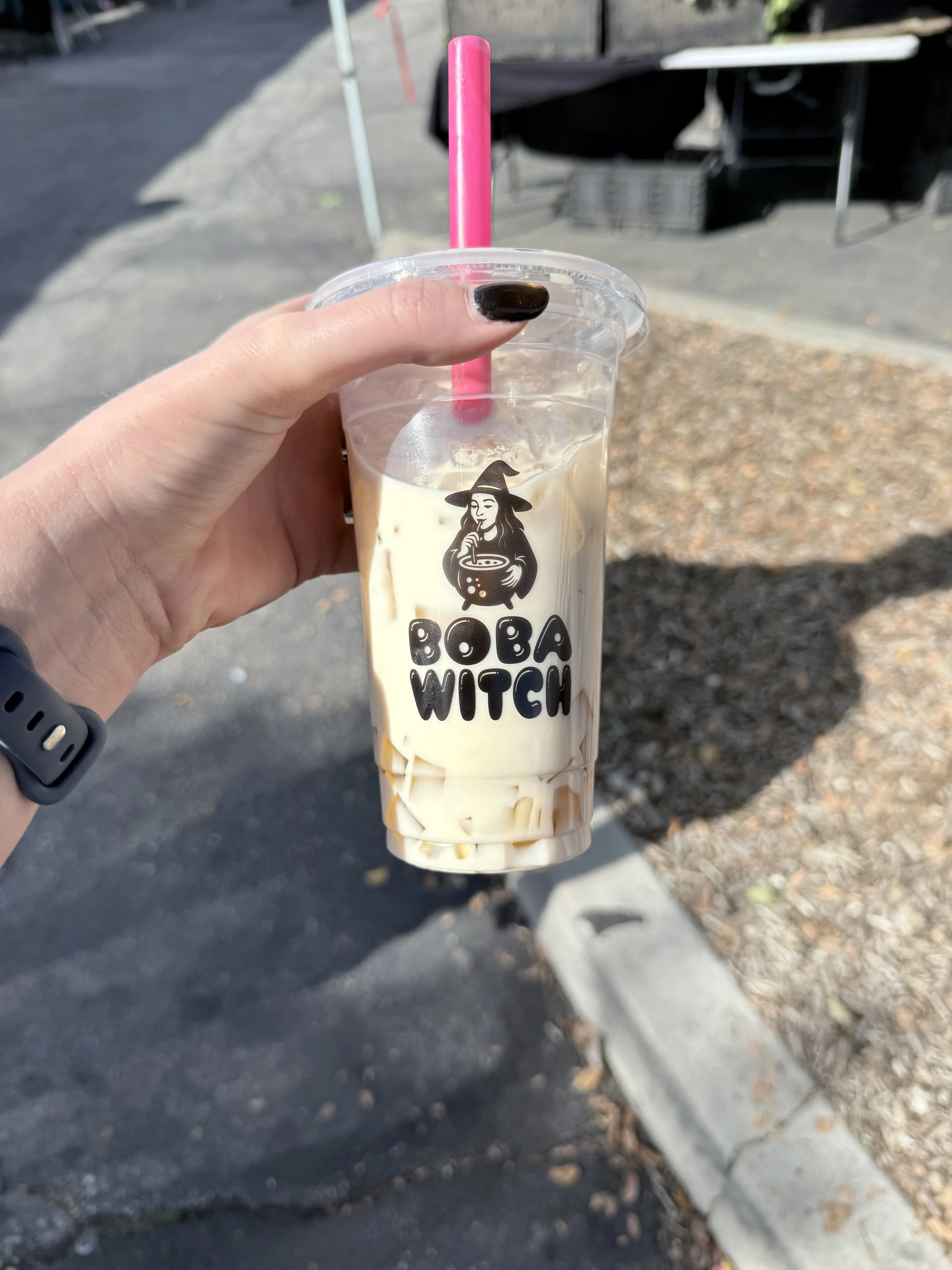 Hand holding a cup of bubble tea with the logo 'BOBA WITCH' on the front, featuring a witch holding a bowl, with a straw in a bright outdoor setting.