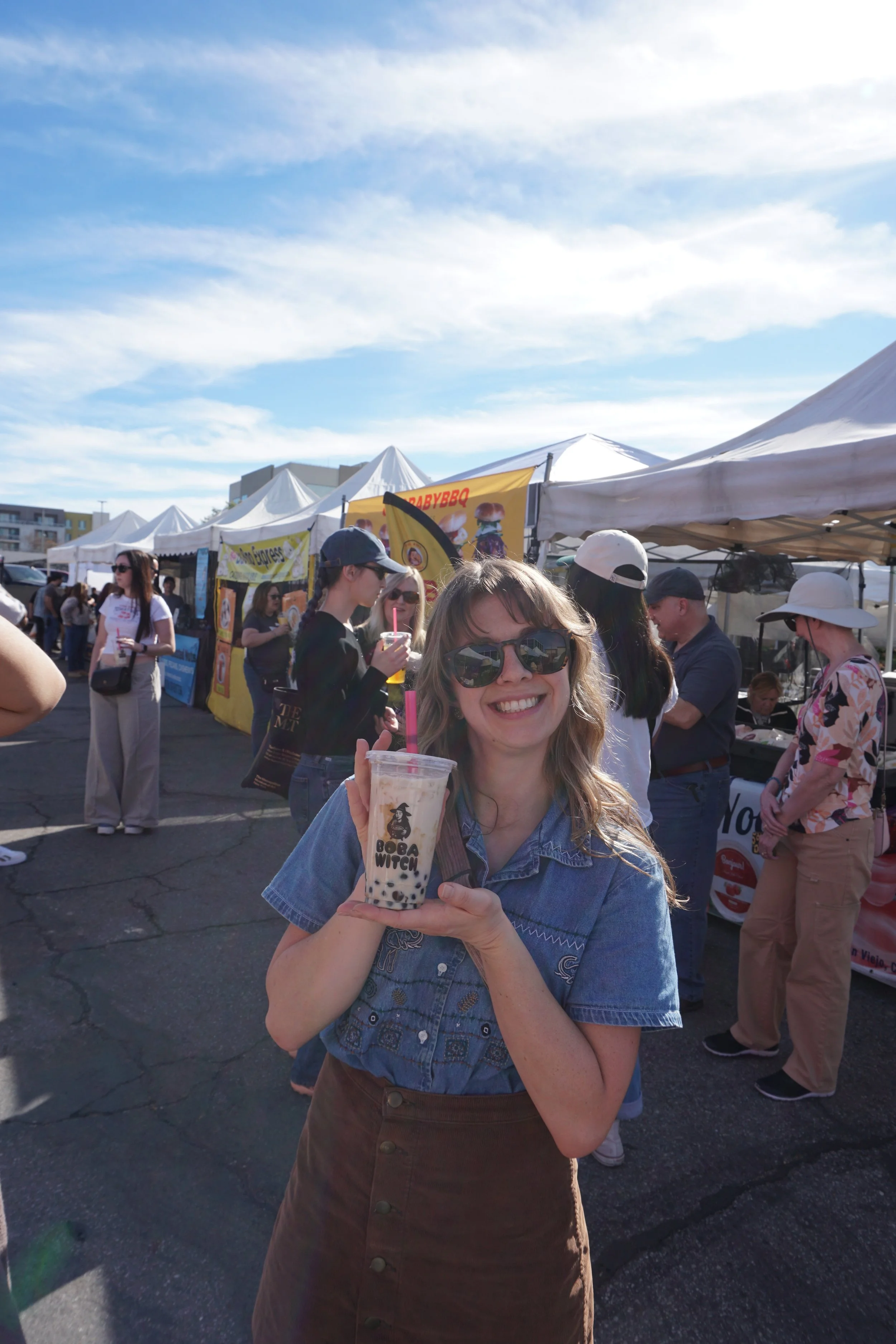 A smiling woman with sunglasses holding a cup of bubble tea at an outdoor market or festival with white tents and people in the background.