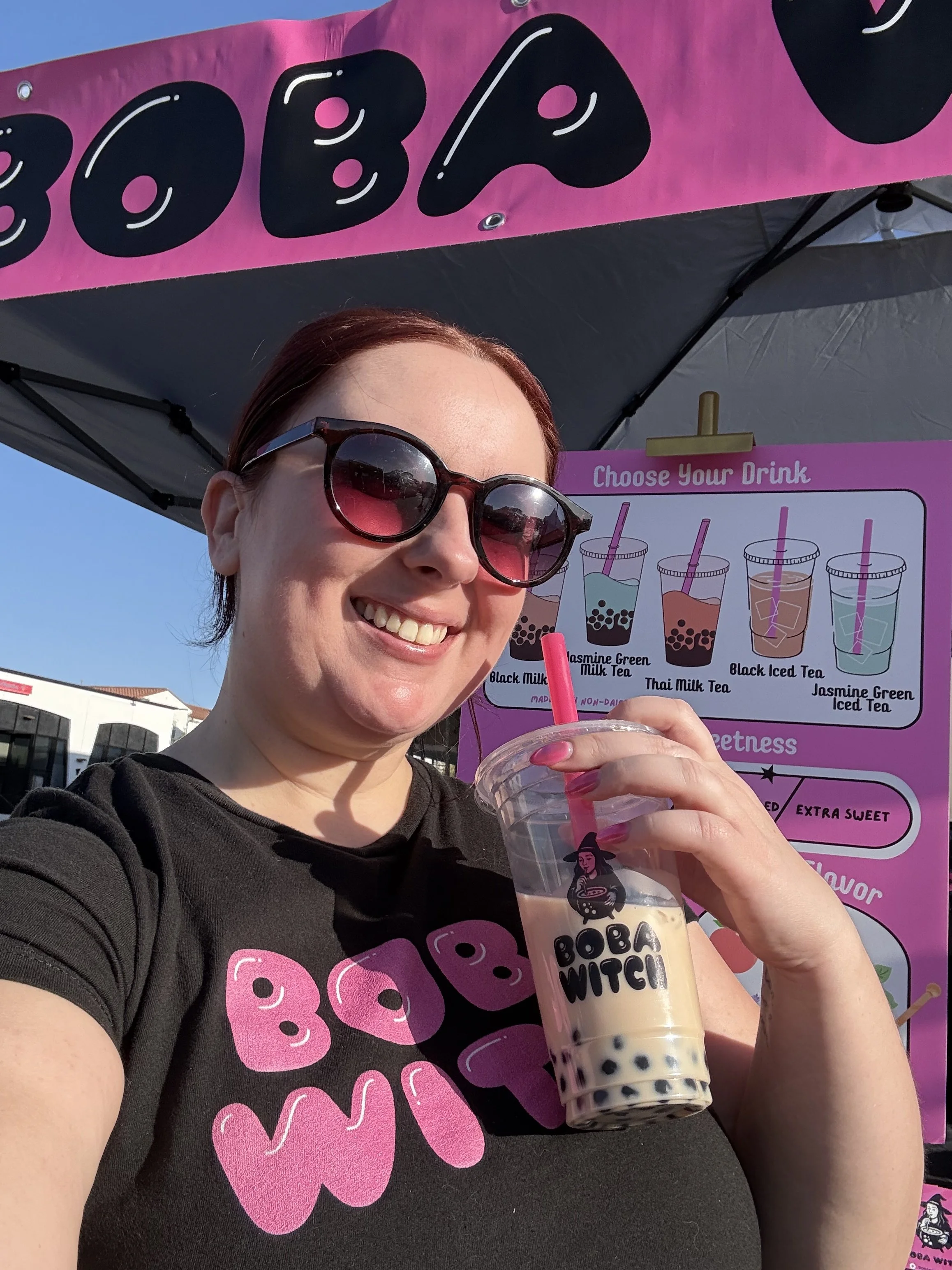 A woman wearing sunglasses and a black Boba Witch T-shirt is holding a bubble tea drink with black tapioca pearls and the Boba Witch logo on the cup. She is smiling and standing in front of a pink booth with bubble tea menu and signage under a black 