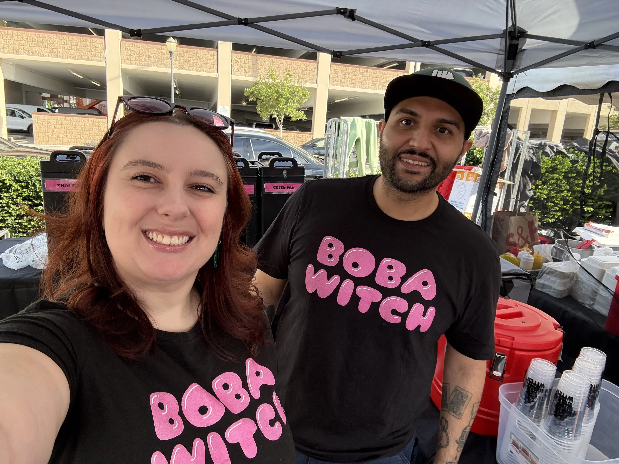 A smiling woman with red hair and sunglasses on her head stands next to a man with a beard, both wearing black T-shirts with pink text that says "BOBA WITCH." They are at an outdoor booth with various supplies and a canopy, with parking and a building in the background.