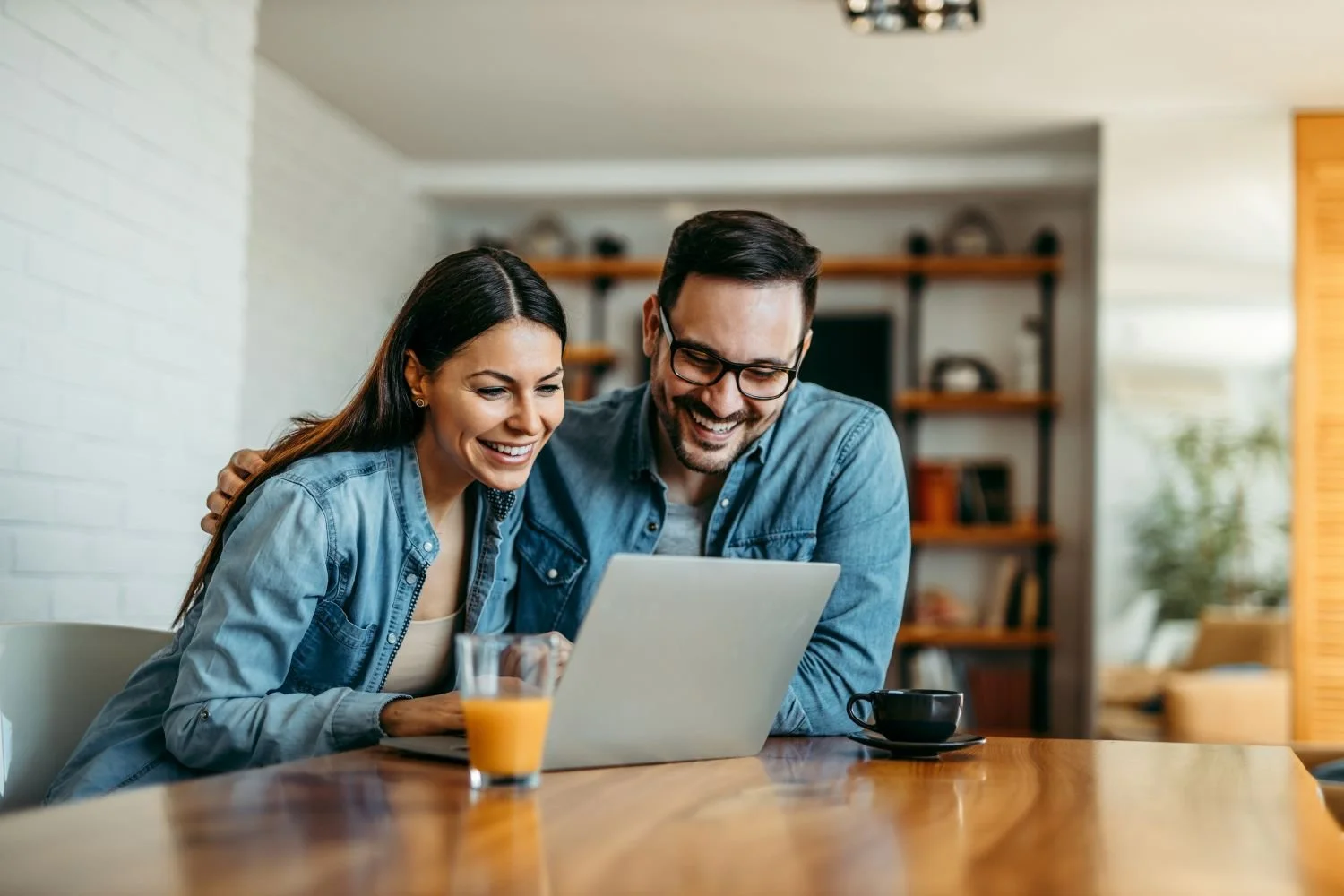 Two people at a home table viewing a laptop together, with coffee and juice nearby.