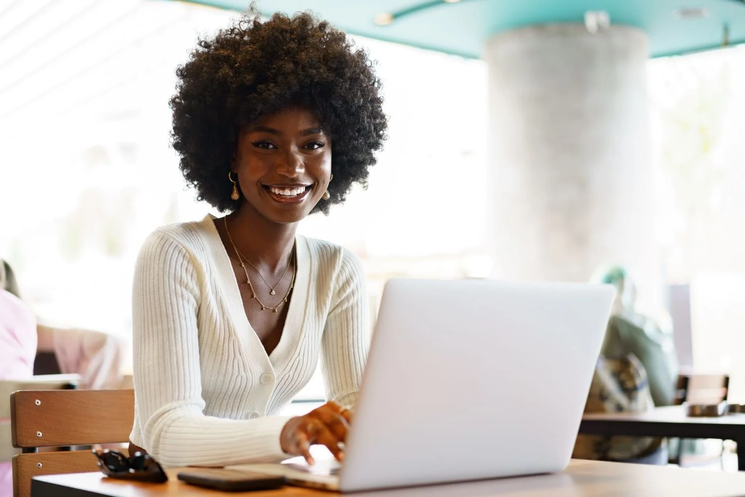 Smiling woman in white top working on a laptop at a café table with phone and sunglasses.