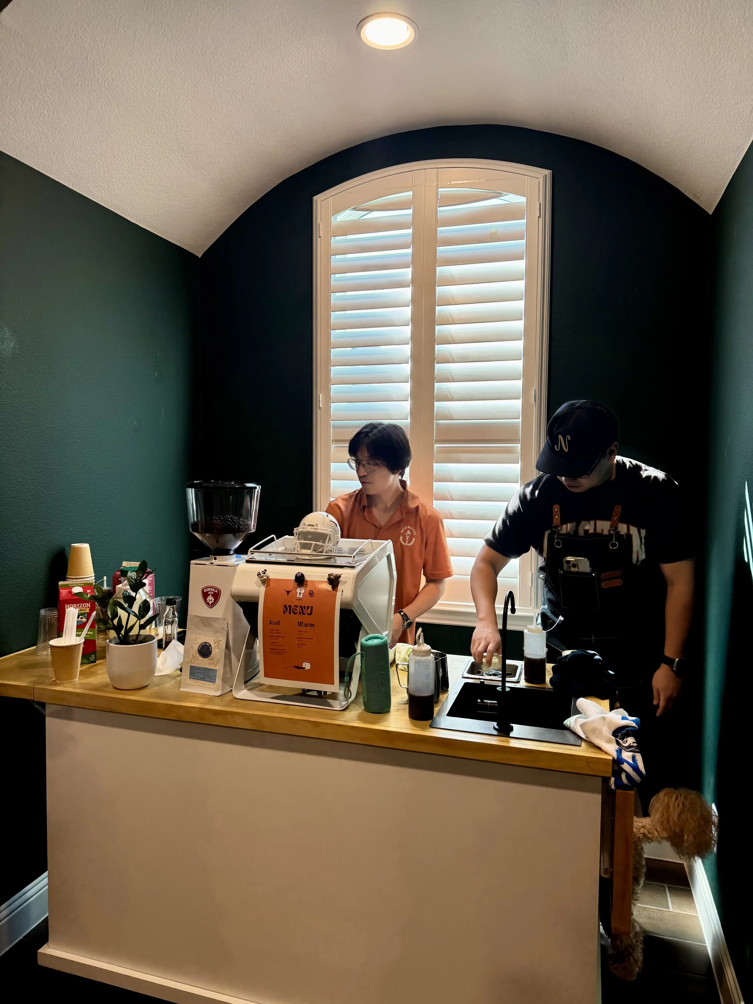 Two people working behind a wooden counter in a small coffee shop, with a dark green wall, a large window with white shutters, and a small dog behind the counter near the floor.