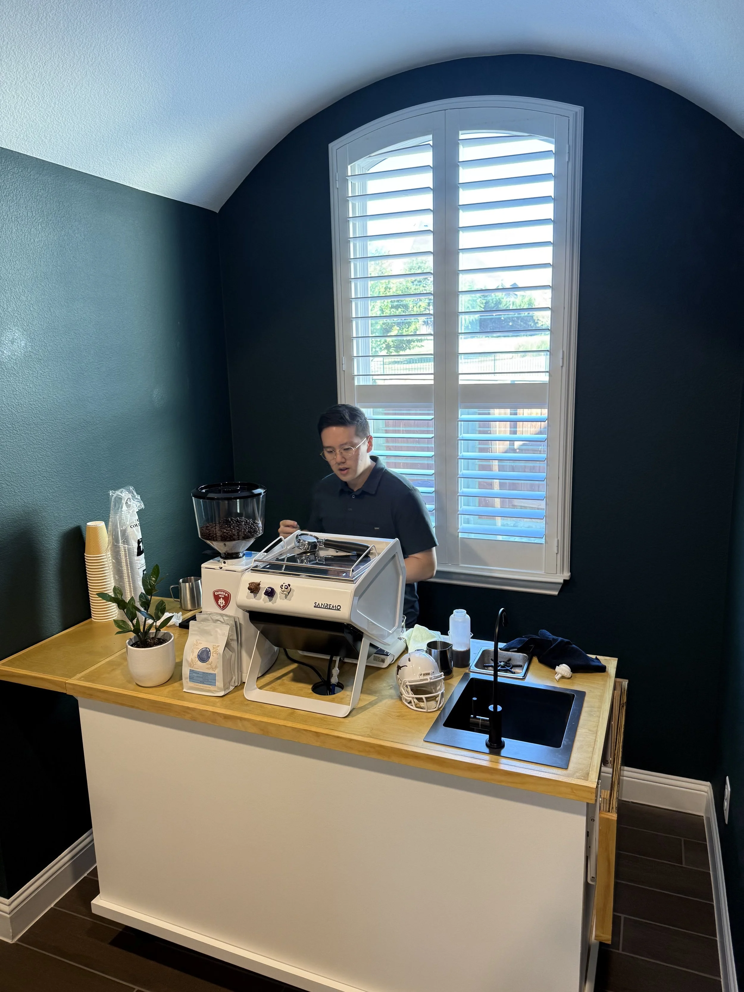 A man making coffee behind a small wooden counter with a coffee machine, a potted plant, a bag of coffee beans, and a helmet, near a window with shutters in a room with dark green walls.