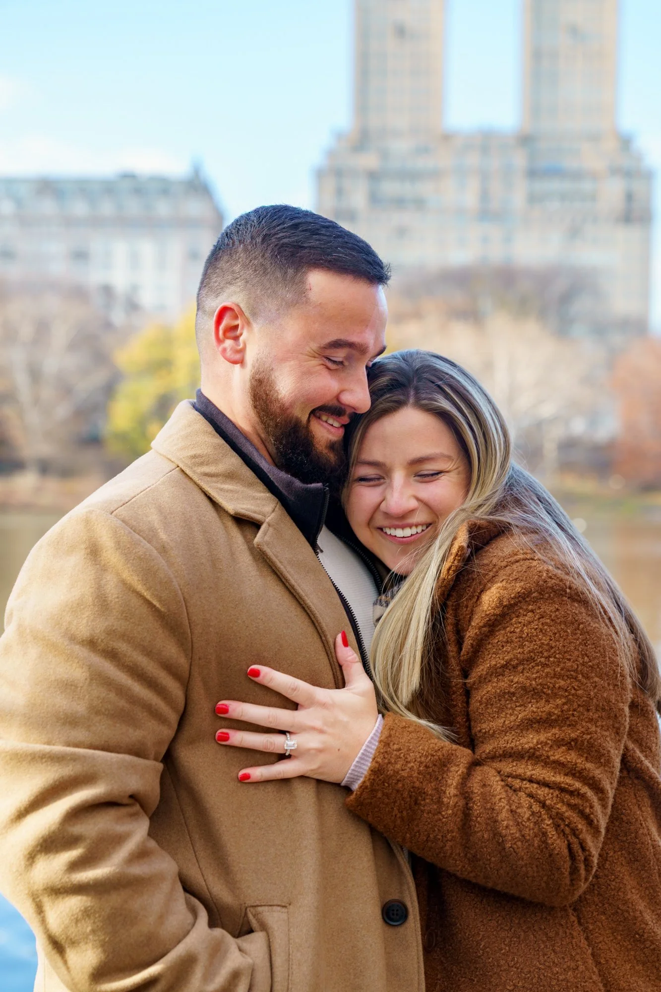 A couple embracing immediately after a proposal in Central Park, with the San Remo Building in the background of the photo