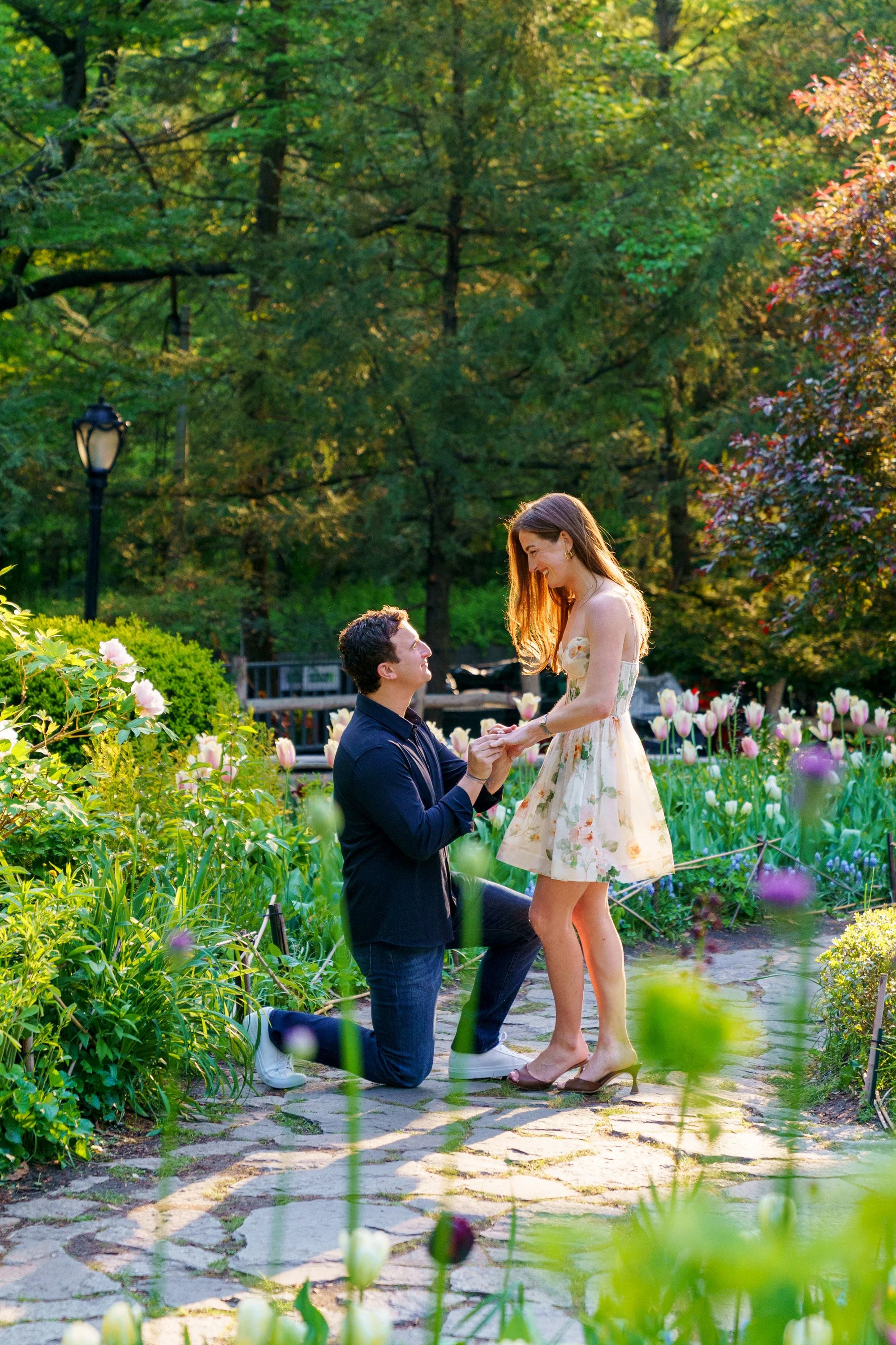 Man on one knee, putting a ring on his fiancee's finger, surrounded by tulips and other blooms in Central Park's Shakespeare Garden
