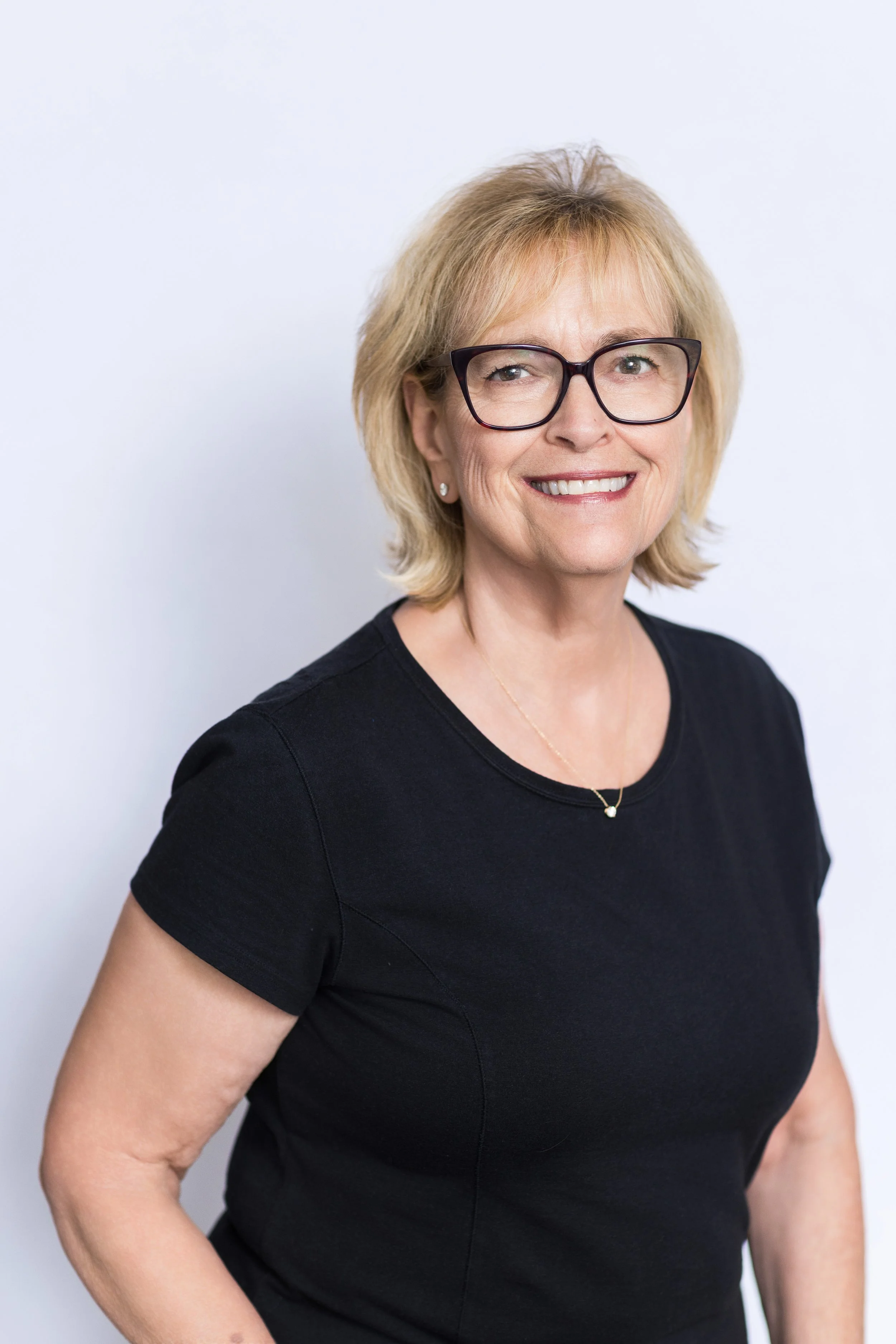 A smiling middle-aged woman with blonde hair, wearing glasses, a black shirt, and earrings, standing against a plain white background.