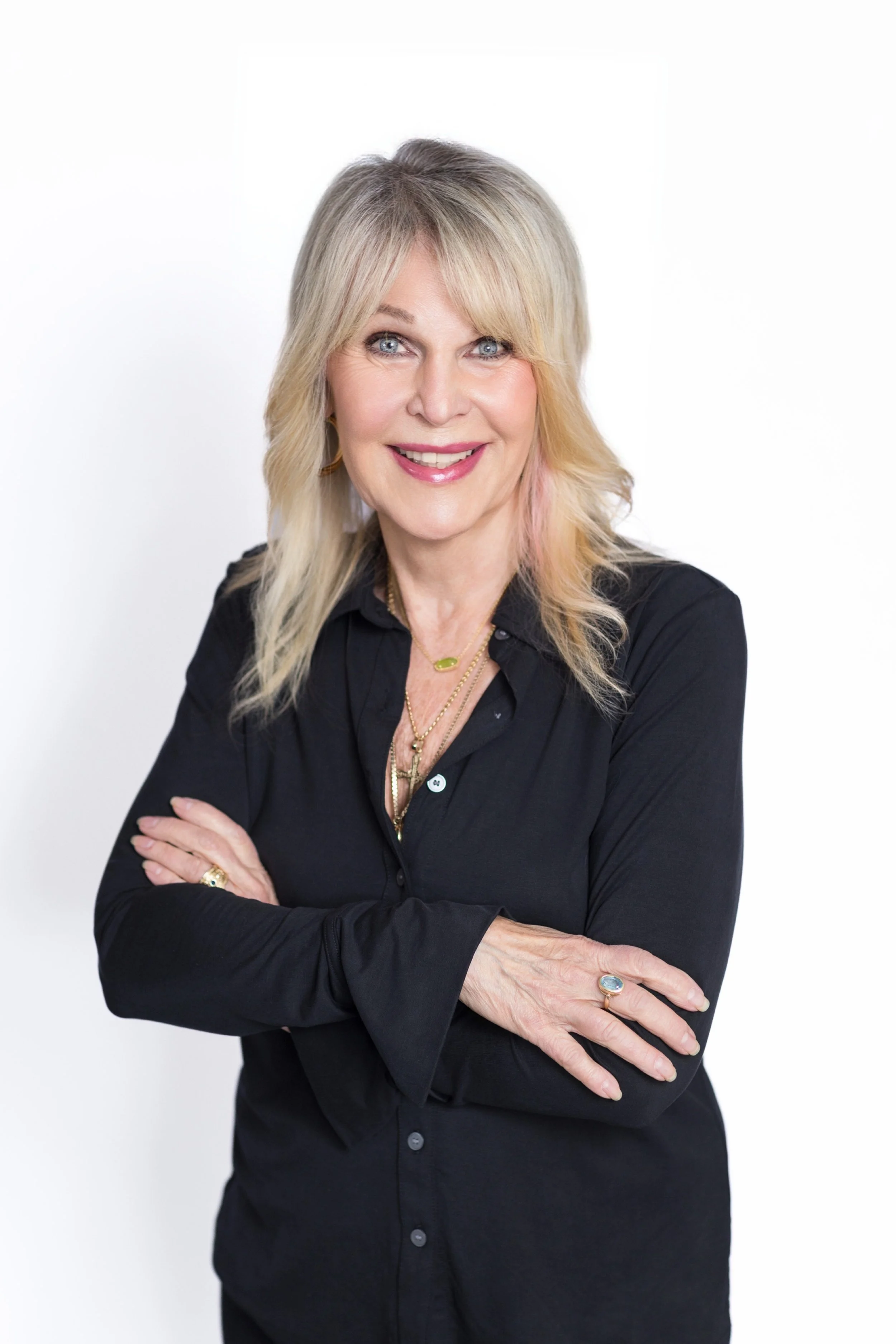 A woman with blonde hair and blue eyes smiling, wearing a black shirt and gold jewelry, standing with arms crossed against a white background.