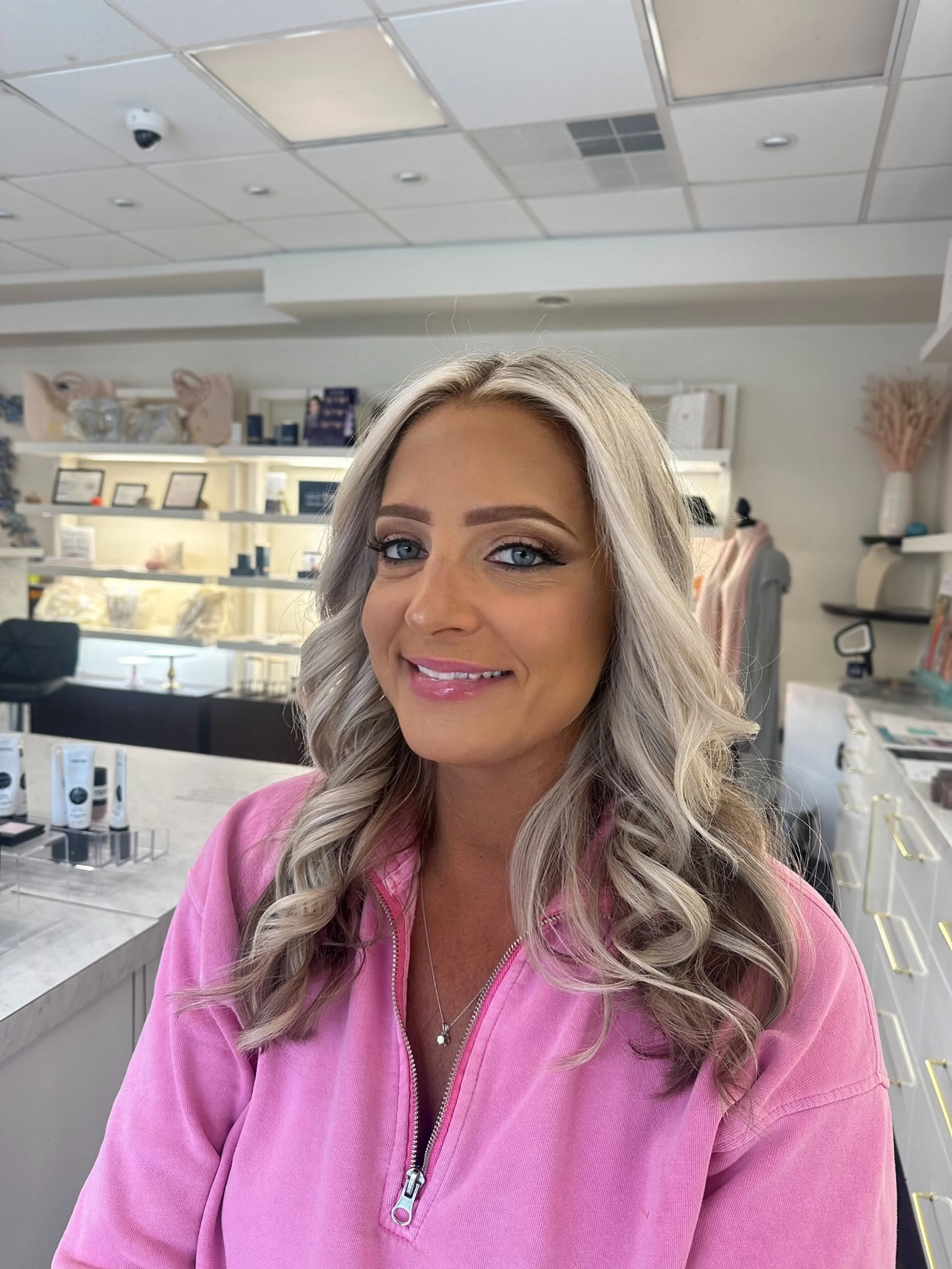 A woman with blonde, wavy hair and blue eyes smiling at the camera inside a store that sells home decor and accessories. She is wearing a pink zip-up top and a necklace with a small pendant.