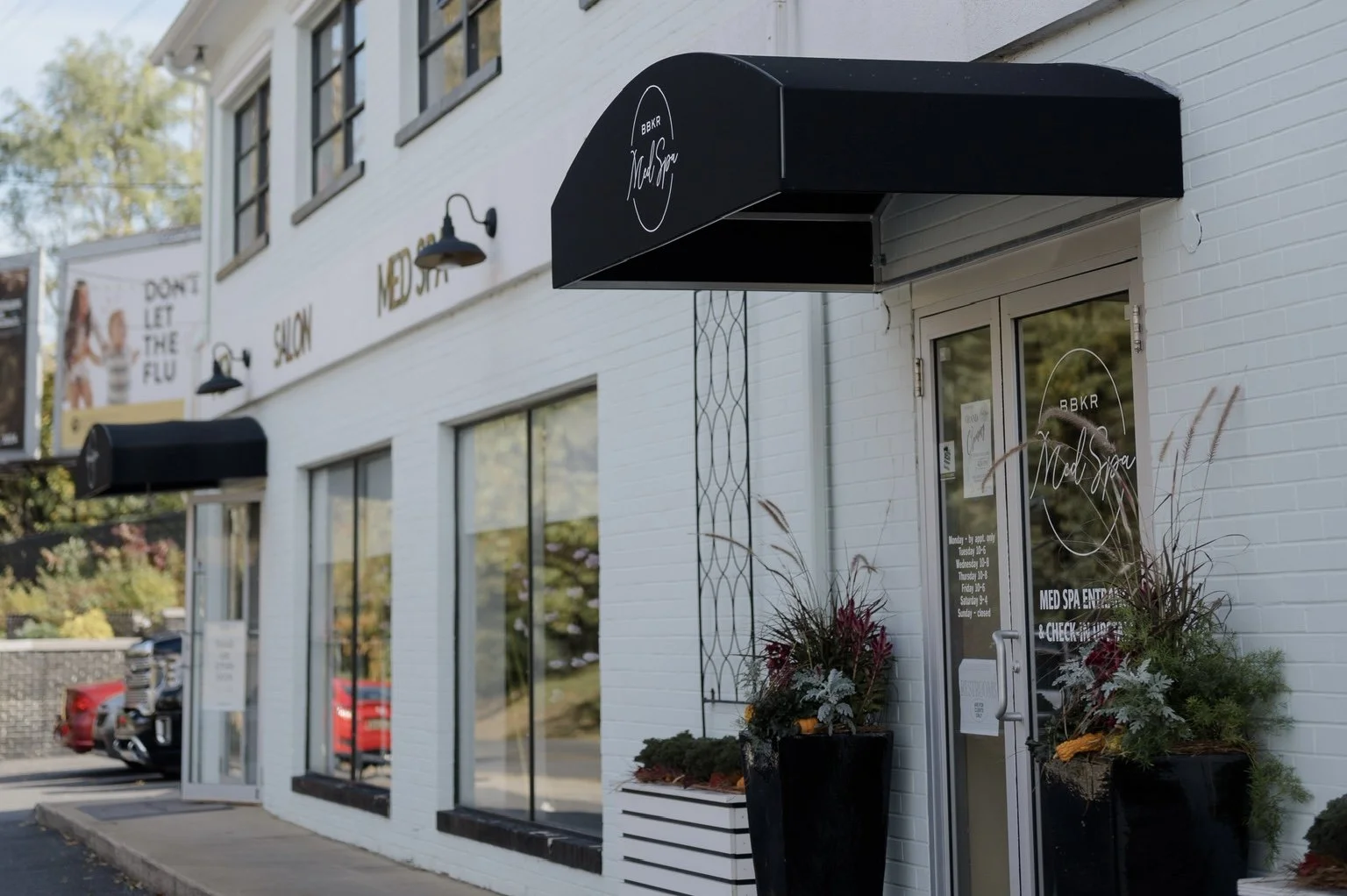 Exterior of a modern white brick spa building with black awnings, potted plants, and glass doors, featuring signs and decor for a med spa.