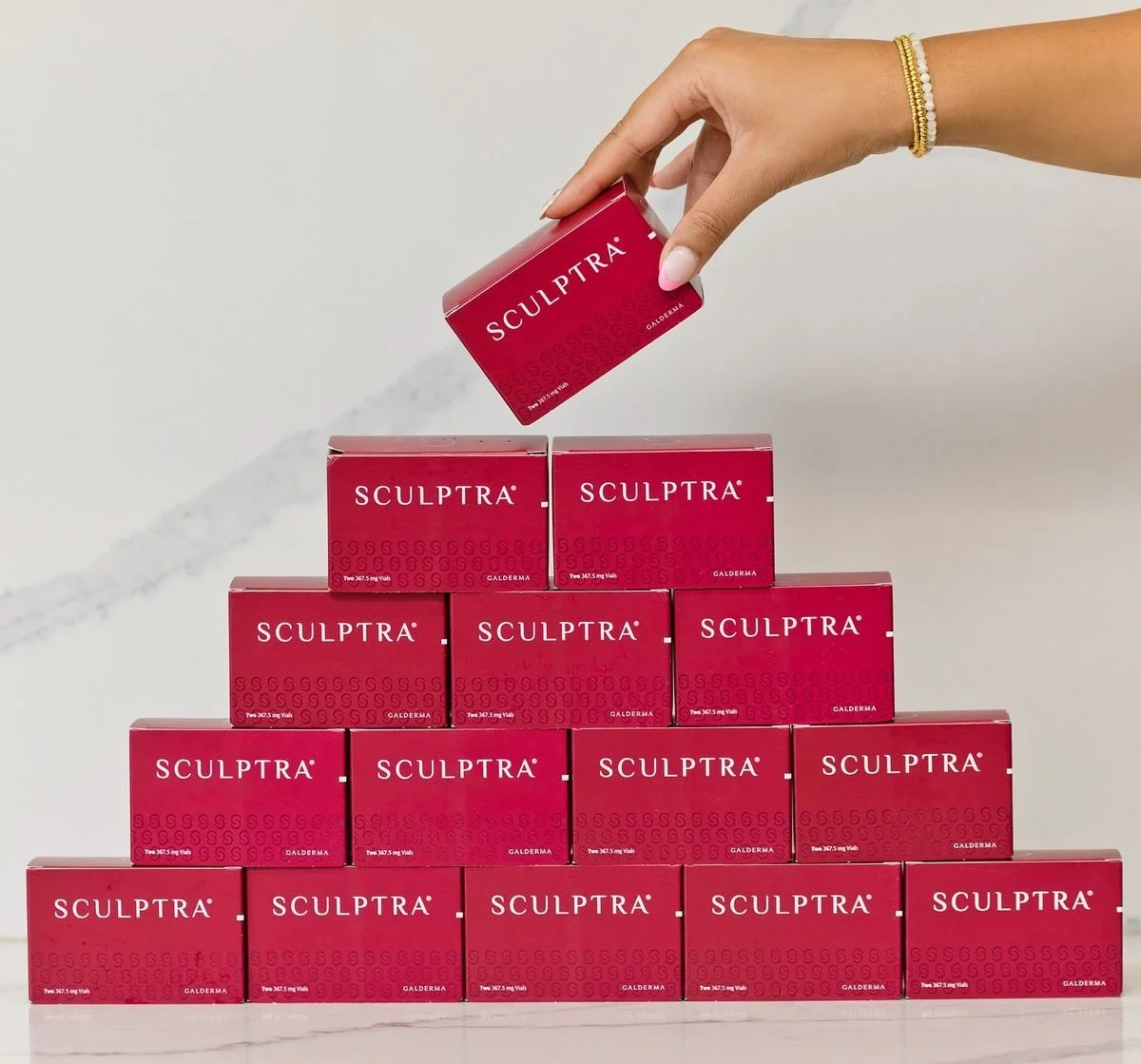 A hand stacking red boxes of Sculptra in a pyramid formation against a gray background. The person is wearing a gold bracelet.