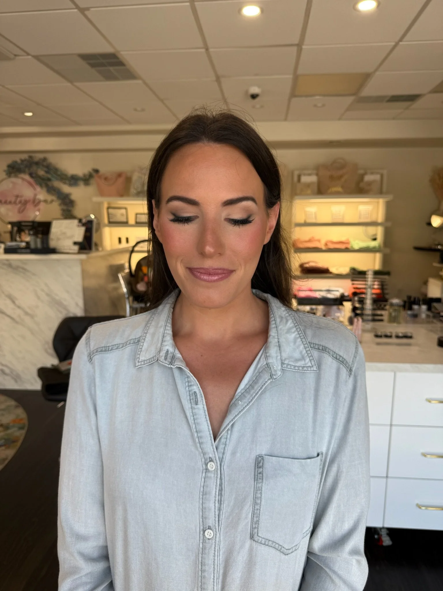 A woman with brown hair and makeup, wearing a light denim shirt, stands indoors with her eyes closed and a slight smile in a well-lit store or cafe.
