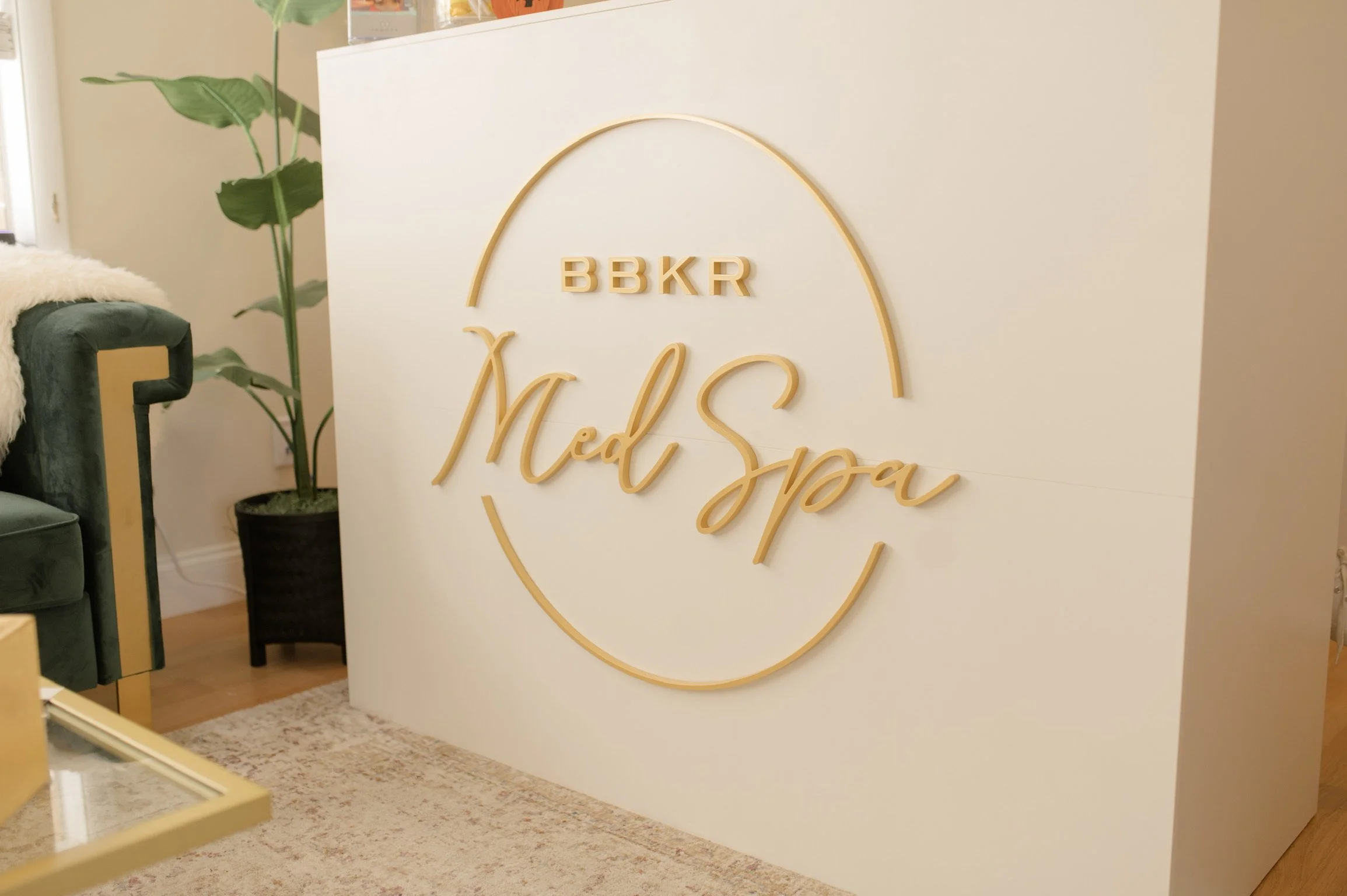 White reception desk with gold sign reading "BBKR Med Spa" and a decorative oval outline, located in a modern spa reception area with a green velvet chair, a potted plant, and plush beige rug.