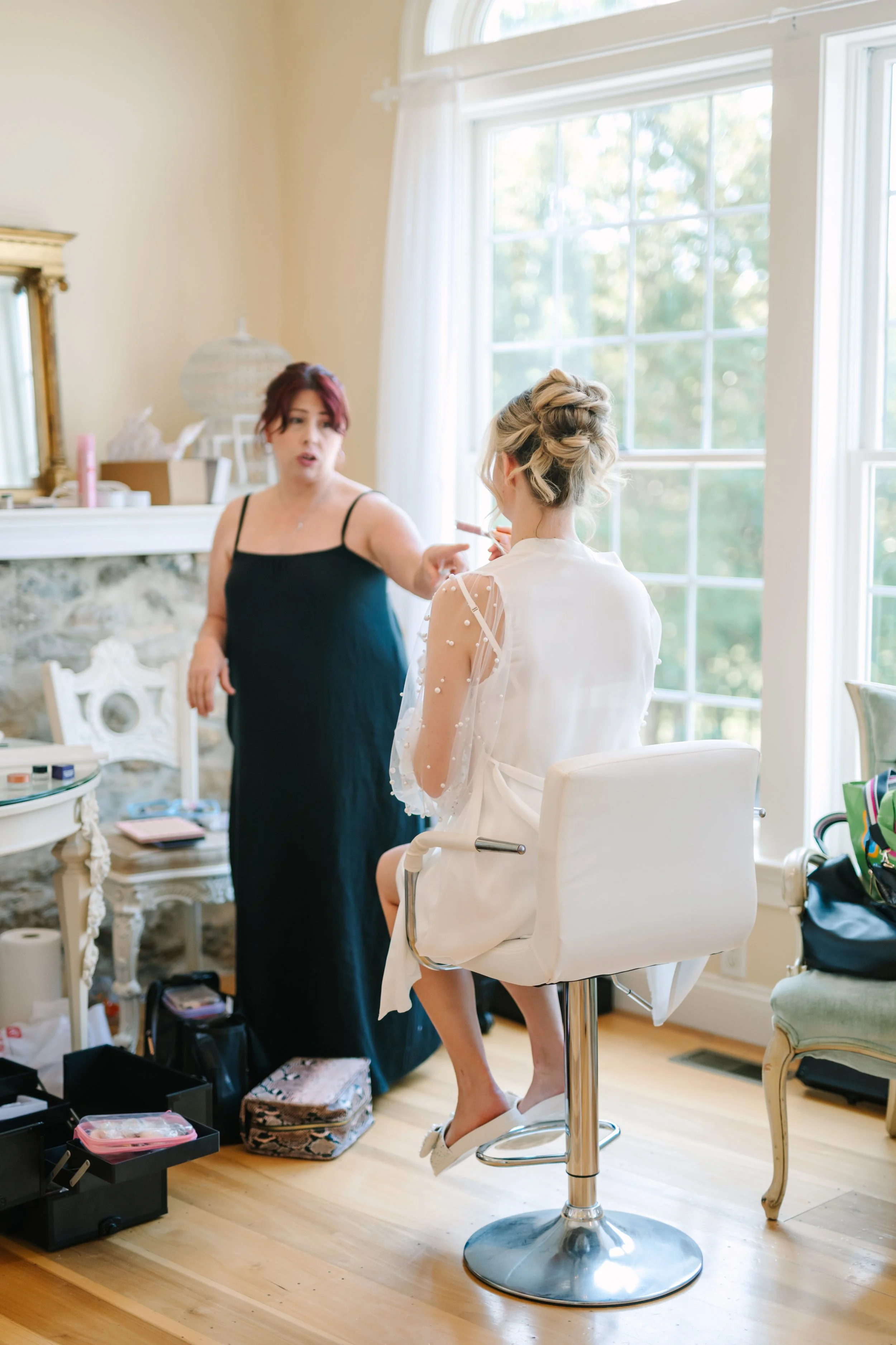 A bride sitting on a white makeup chair getting her makeup done by a makeup artist in a brightly lit room with large windows.