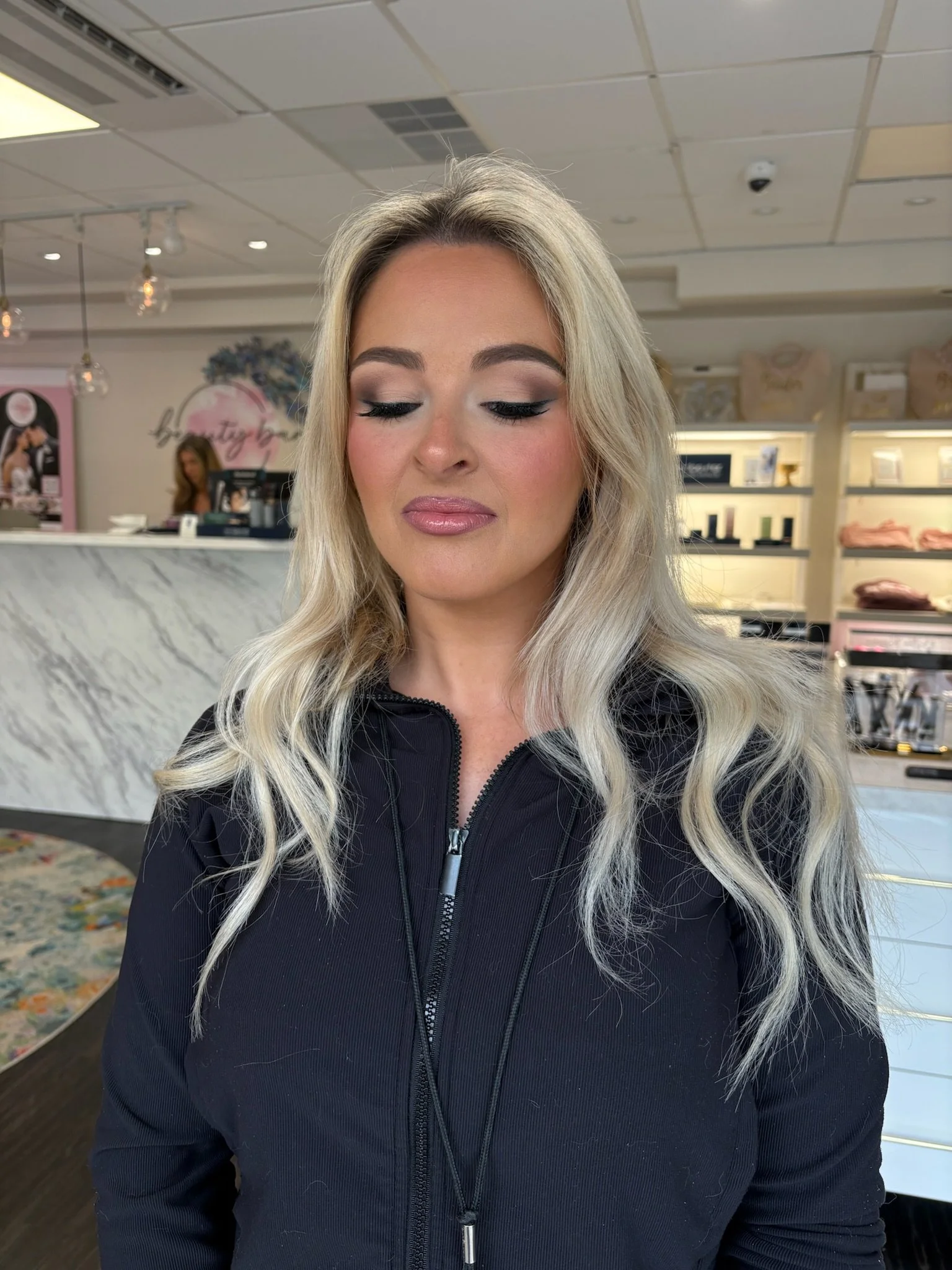 A woman with long blonde wavy hair, makeup, and wearing a black zip-up jacket, standing inside a store with a marble counter and display shelves in the background.