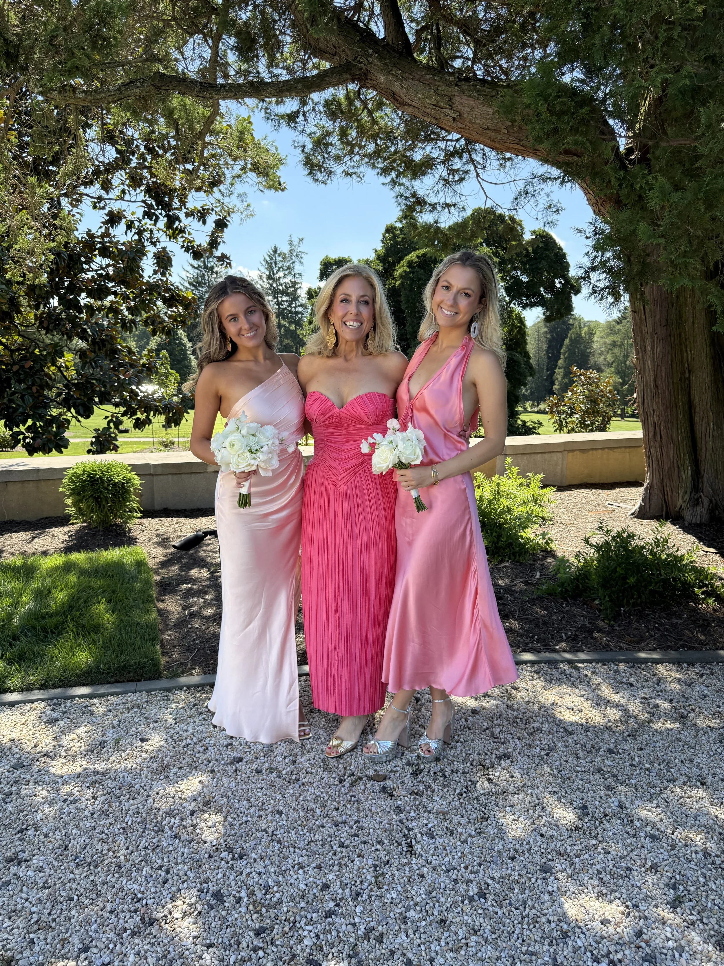 Three women in pink dresses standing outdoors under a large tree, holding white bouquets, smiling at the camera on a sunny day.