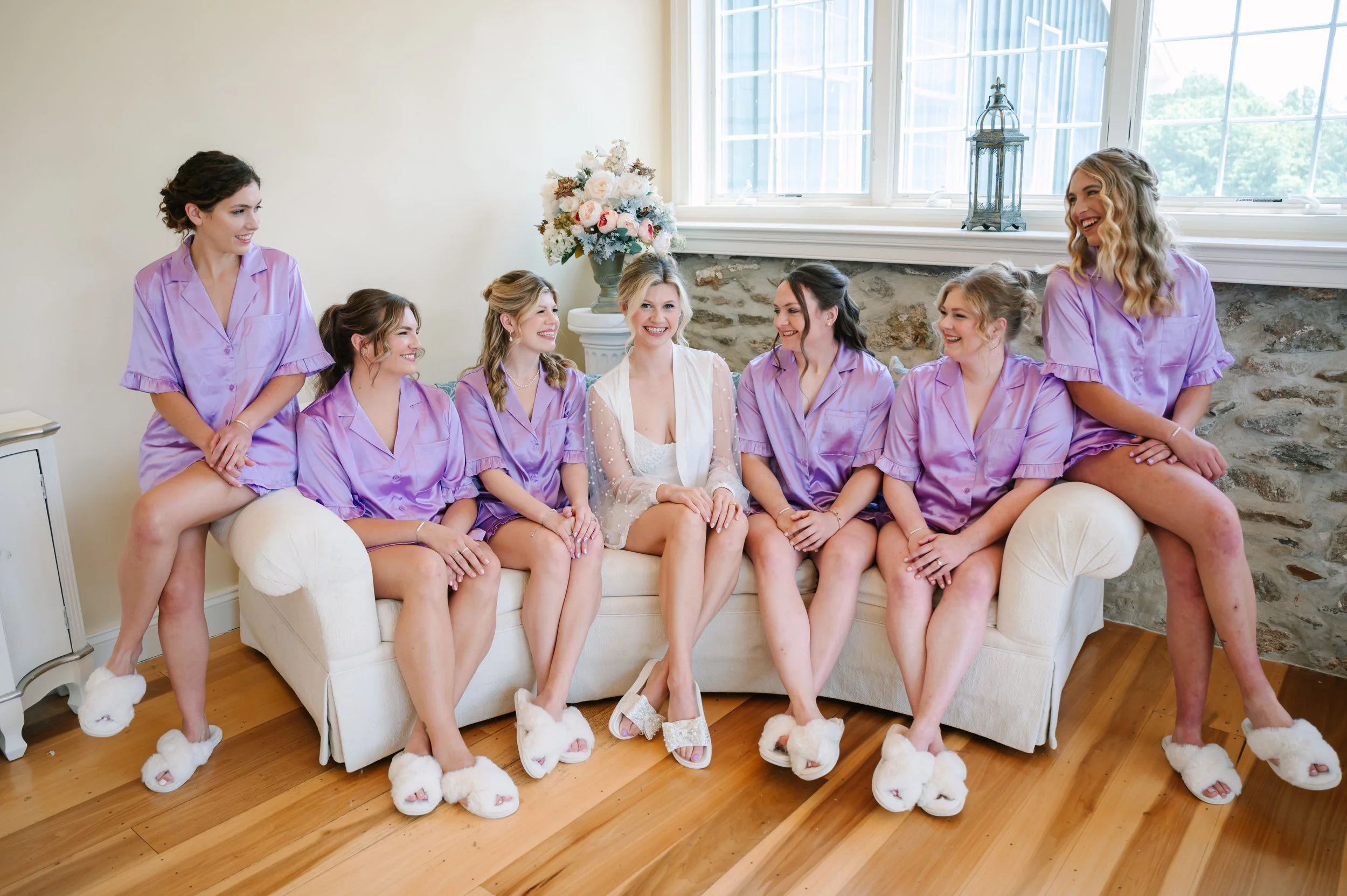 A bride and six bridesmaids sitting on a couch in a room with a stone wall. They are all wearing lavender pajamas and bunny slippers, smiling and looking at each other.