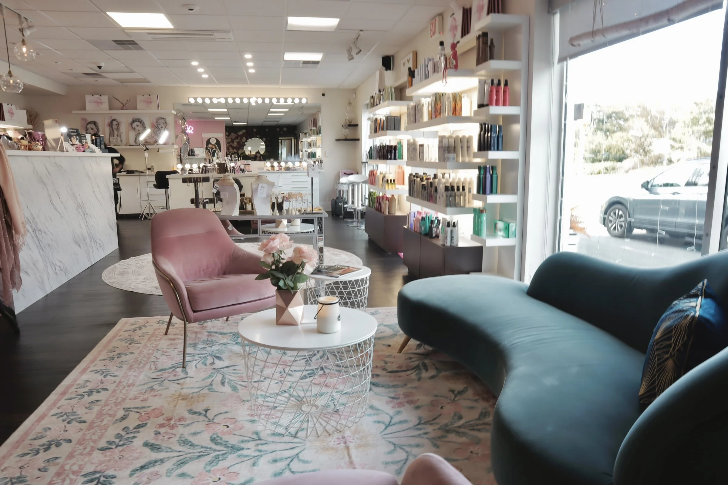 Interior of a boutique shop with pink and teal seating, floral rug, white circular coffee table, and shelves stocked with beauty or skincare products near a large window.