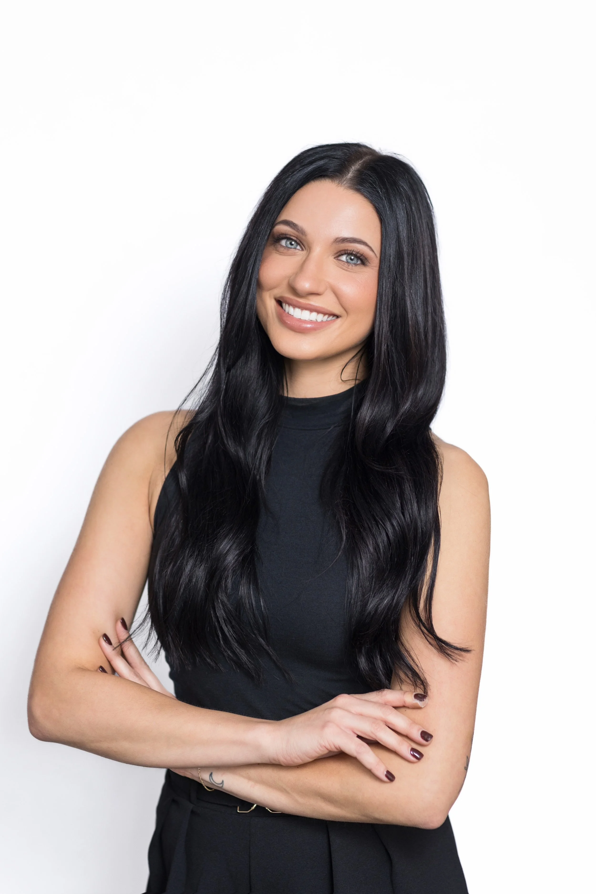 A young woman with long black hair, blue eyes, and fair skin, smiling, wearing a sleeveless black top, standing against a plain white background.