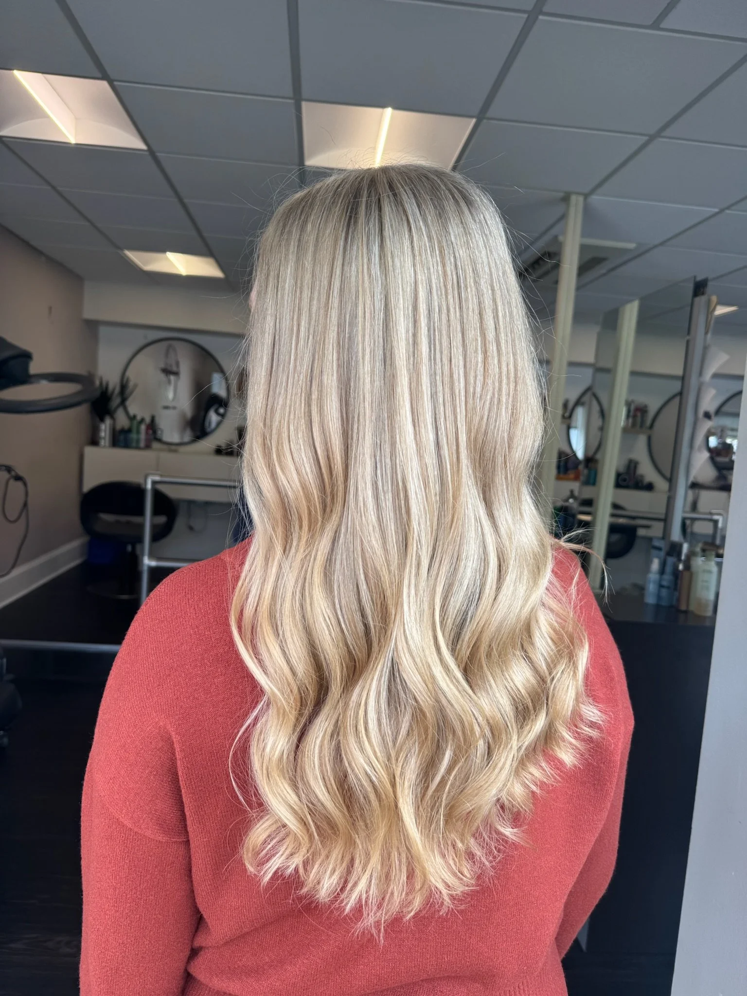 Back view of person with long, wavy blonde hair wearing a red top inside a salon.