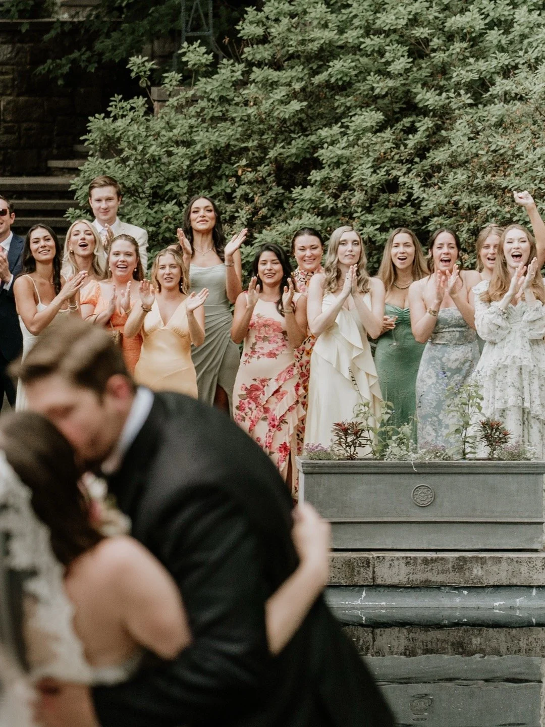 A couple kissing during their wedding, with a group of friends and family celebrating in the background outdoors.