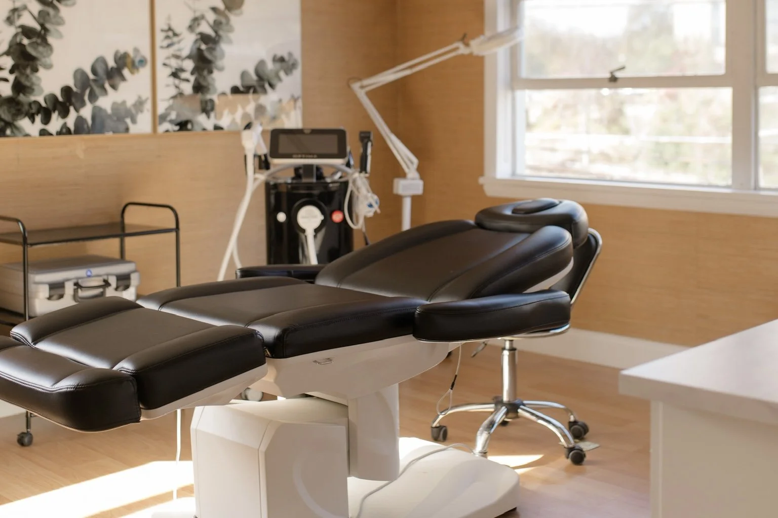 Medical examination or treatment room with adjustable black leather chair, medical equipment, and large window with outside view.