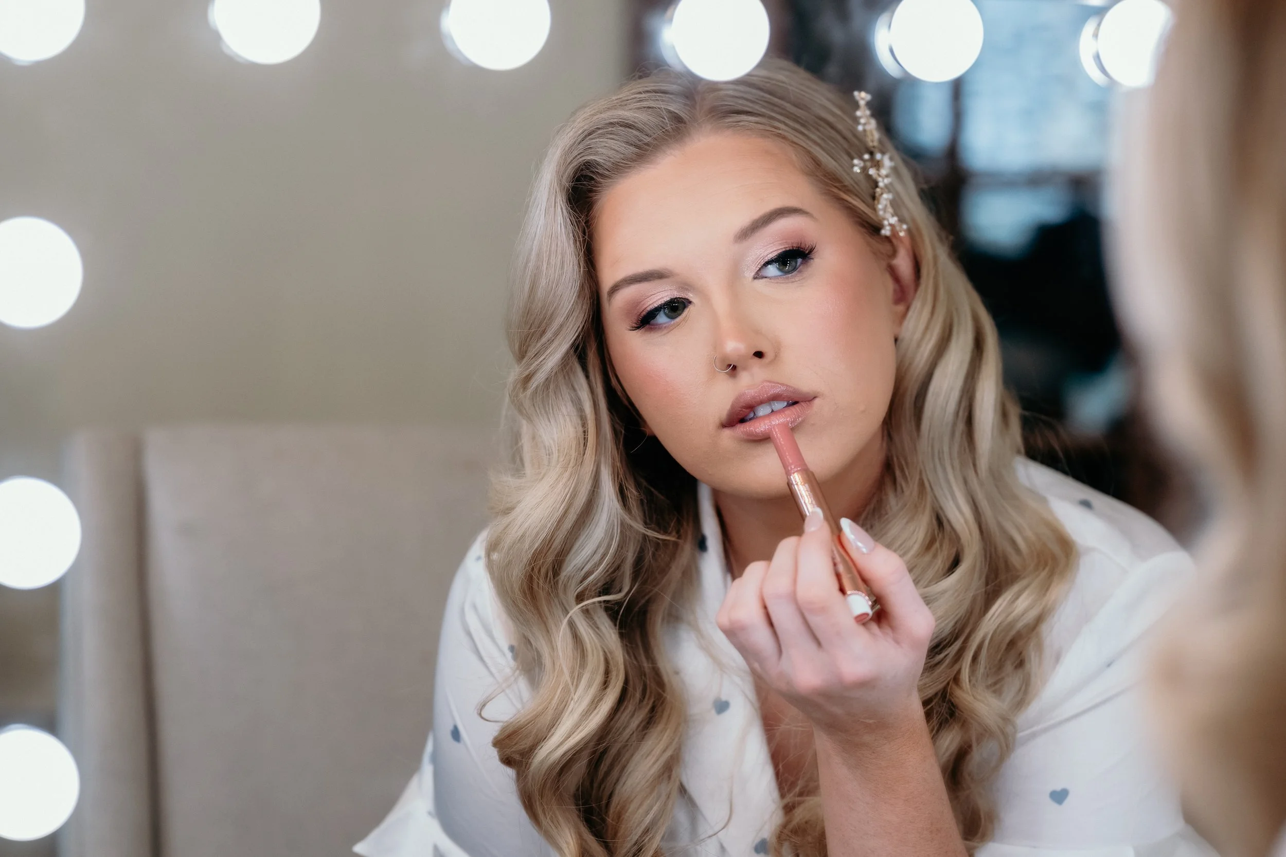 A young woman with blonde, curly hair applying lipstick in front of a mirror with bright lights, wearing a white top with small blue hearts and a decorative hairpin.