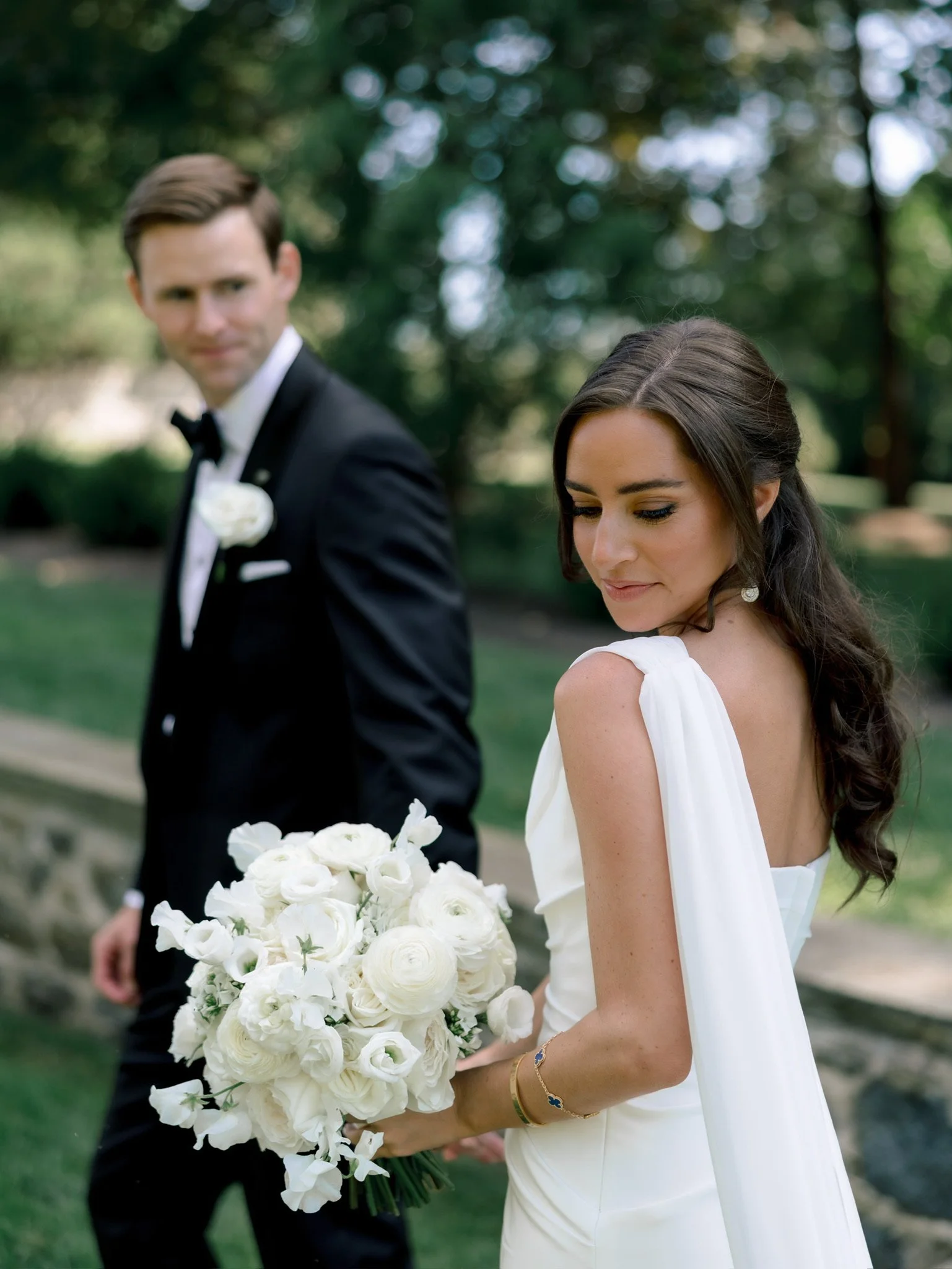 A bride holding a bouquet of white flowers with a groom in a tuxedo standing in the background at an outdoor wedding.