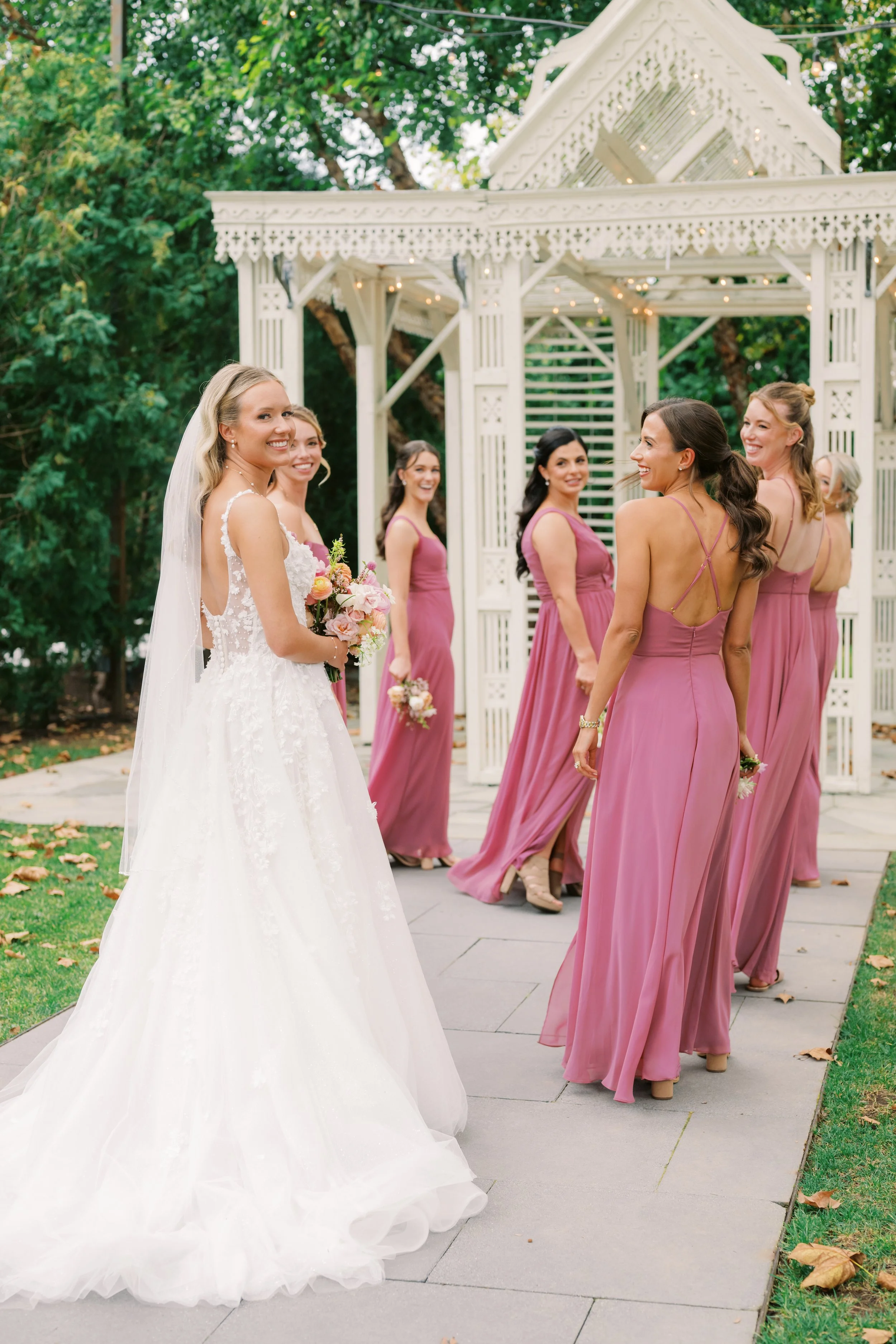 A bride and her bridesmaids standing outside near a white gazebo, with green trees and fallen leaves in the background. The bride is holding a bouquet and wearing a white lace wedding gown, while the bridesmaids are dressed in mauve dresses and holding smaller bouquets, all smiling and looking at each other.