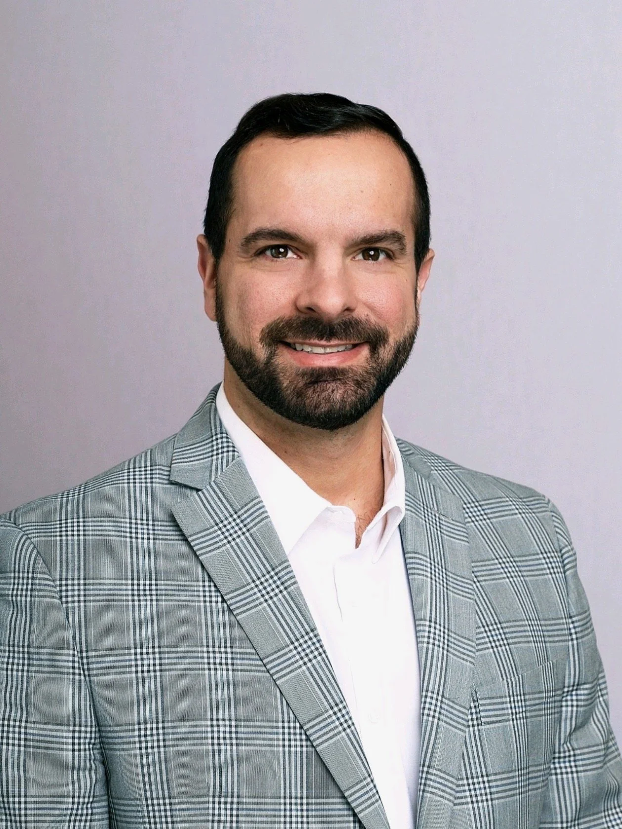 Professional headshot of a man with dark hair and beard, wearing a gray plaid suit and white shirt, smiling against a plain gray background.