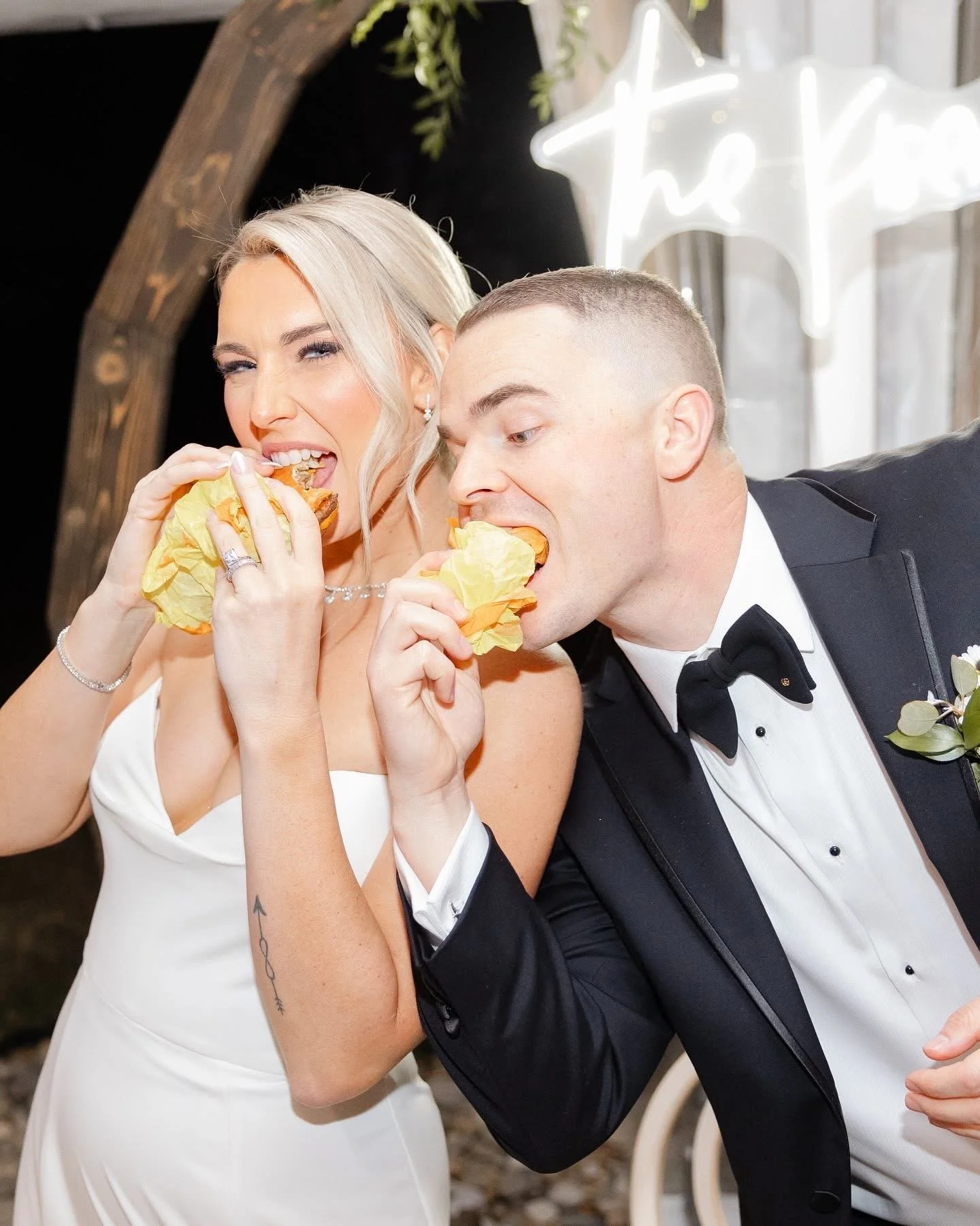 A newlywed couple dressed in wedding attire eating hot dogs at their wedding celebration.