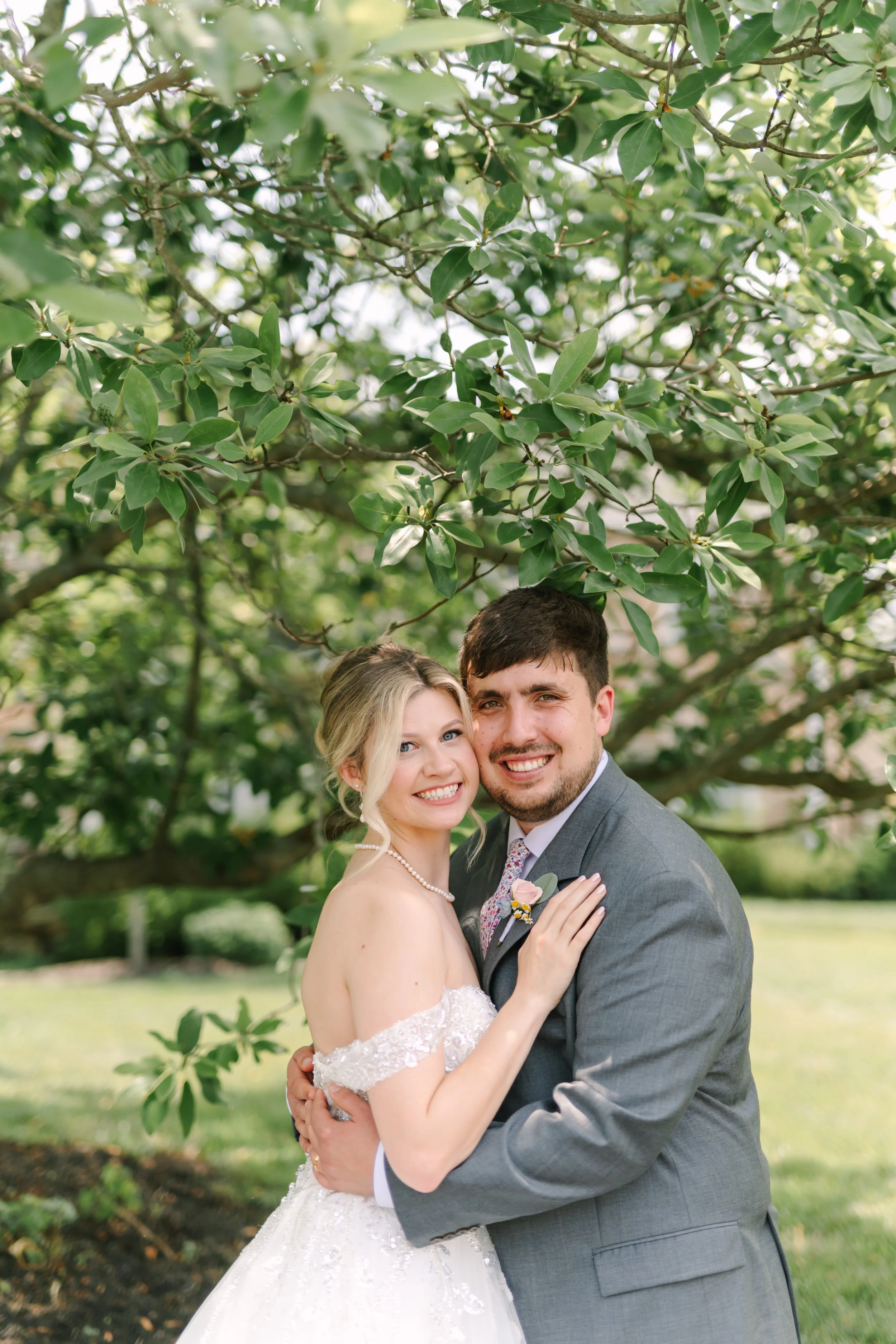 A newlywed couple smiling and embracing outdoors under a large tree, with green leaves and a blurred background.