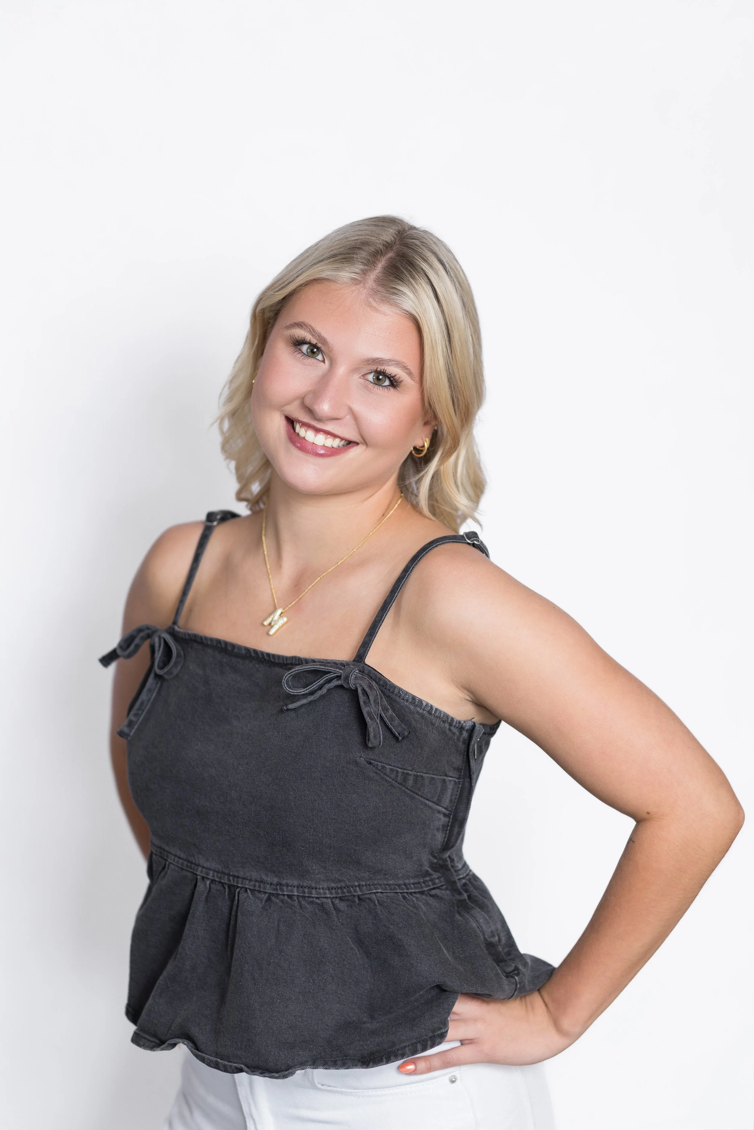 A smiling woman with blonde hair wearing a black sleeveless top with tie straps and white pants standing against a white background.