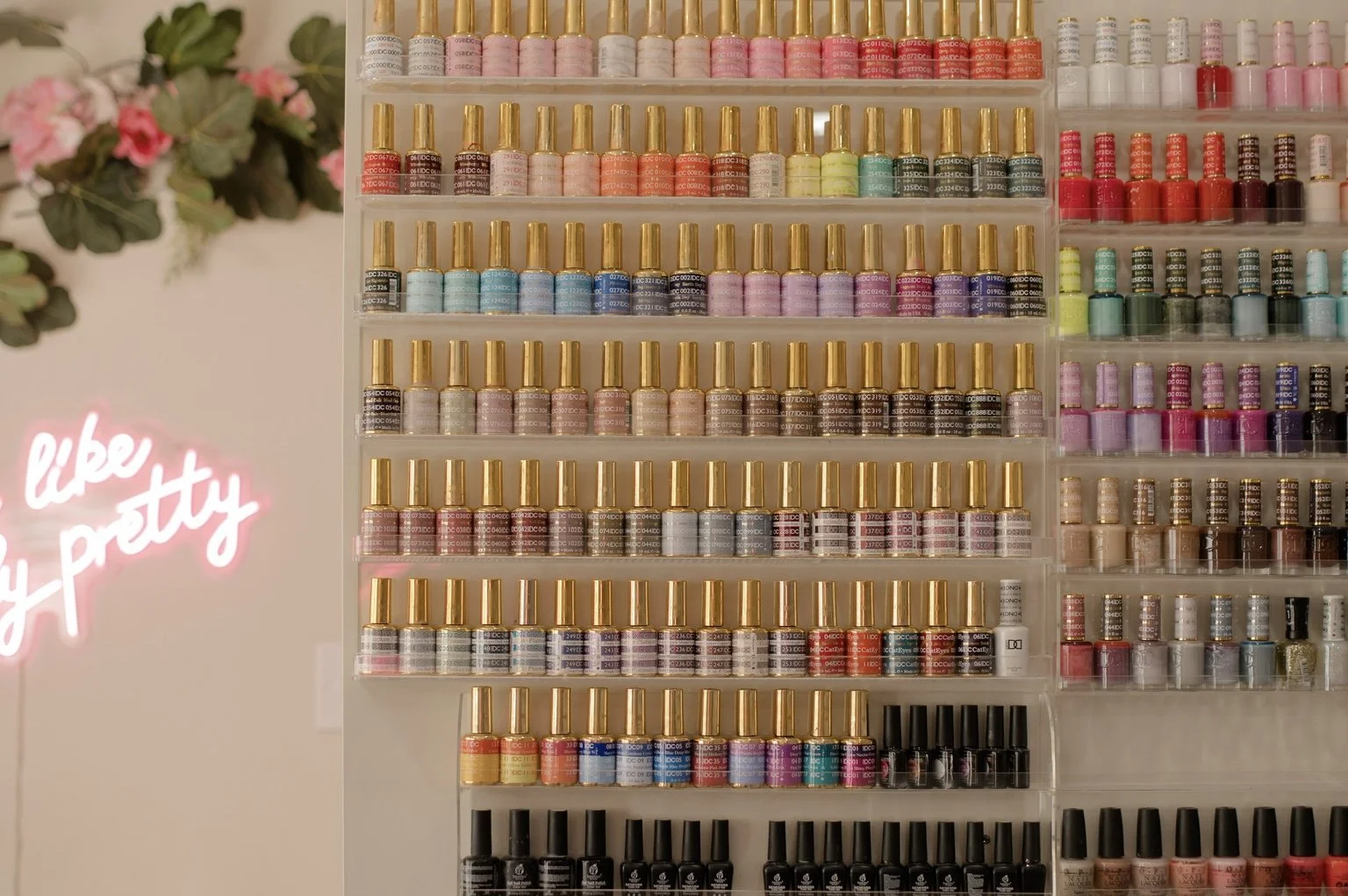 Display shelf filled with various bottles of colorful nail polish, organized in rows by shade, with a pink neon sign reading 'like my pretty' on the left side and some pink flowers in the background.