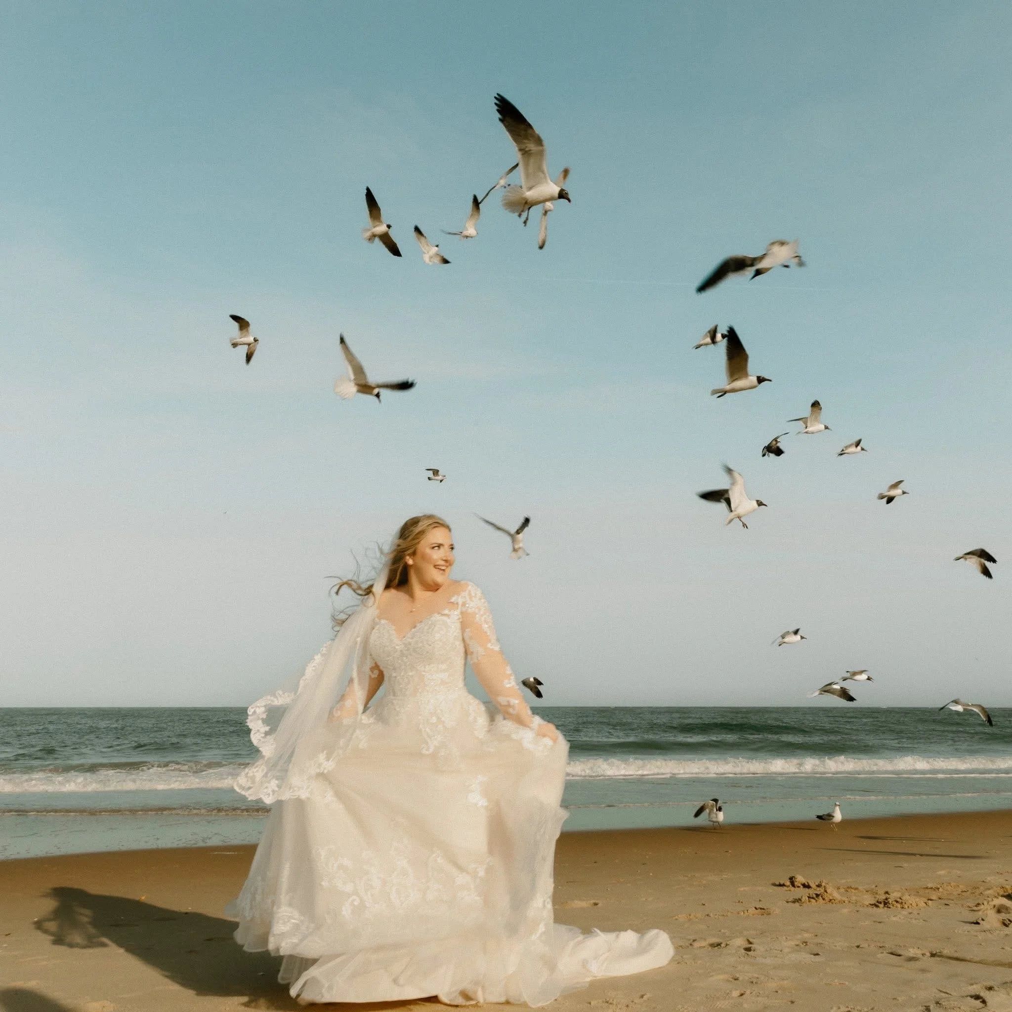 A woman in a lace wedding dress on the beach, smiling, with seagulls flying overhead.
