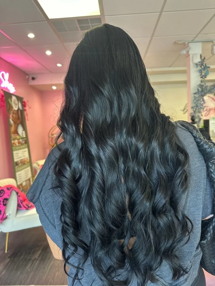 Back view of a woman with long, black, curly hair inside a brightly lit salon or cafe with pink and white decor.