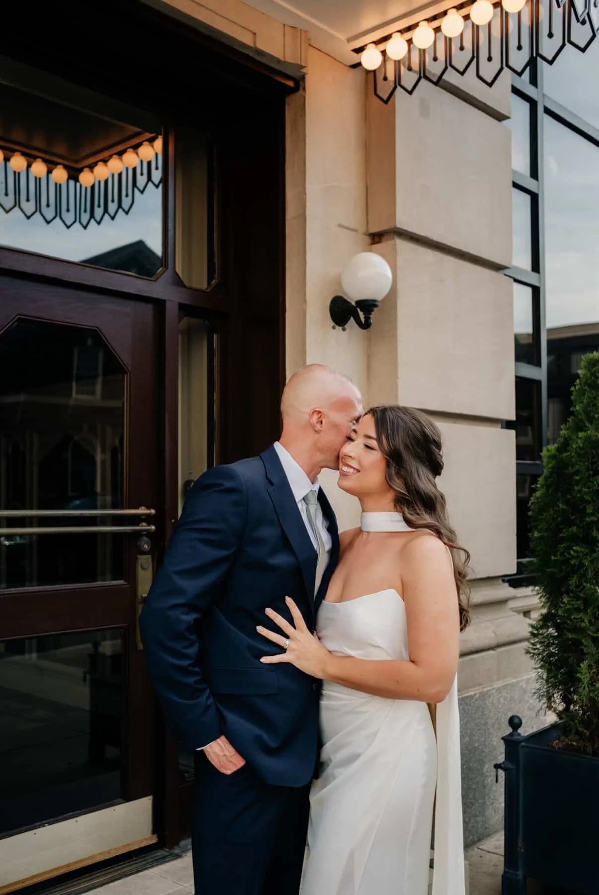 A man in a navy suit kissing a woman in a white dress on the forehead outside a building with a lit string of round lights above.