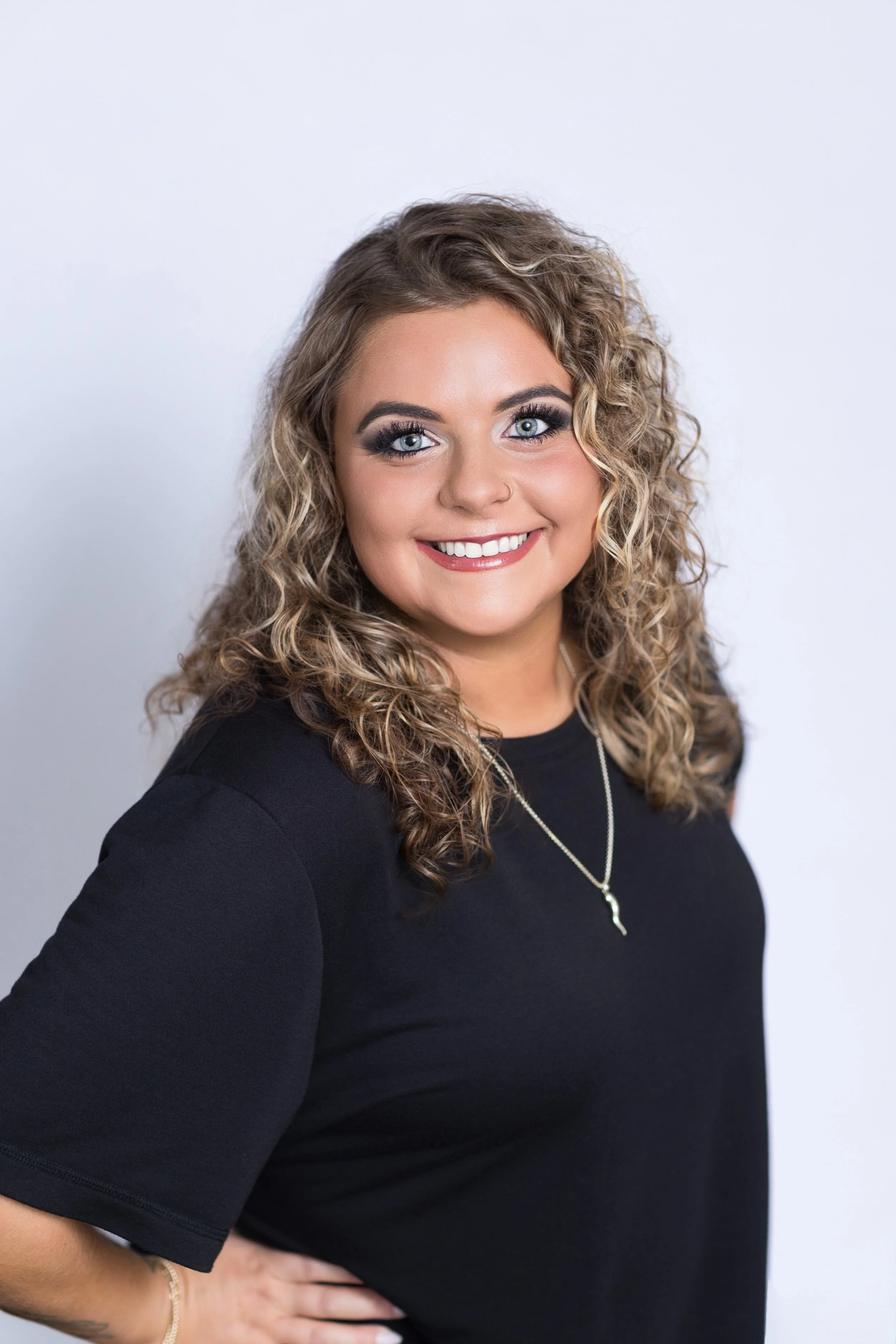 A young woman with curly blonde hair, blue eyes, and makeup, smiling and wearing a black shirt and a silver necklace, standing against a plain white background.