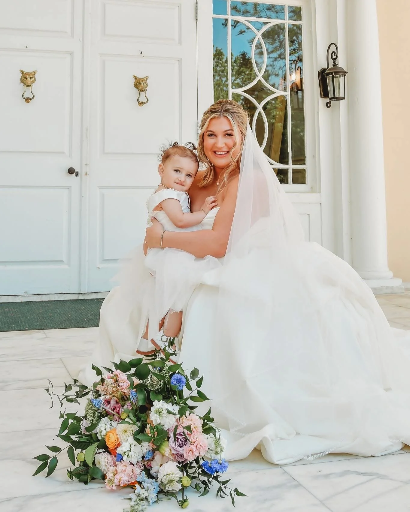 A bride in a white wedding dress sitting outside, holding a young girl in white, with a colorful bouquet of flowers on the ground in front.