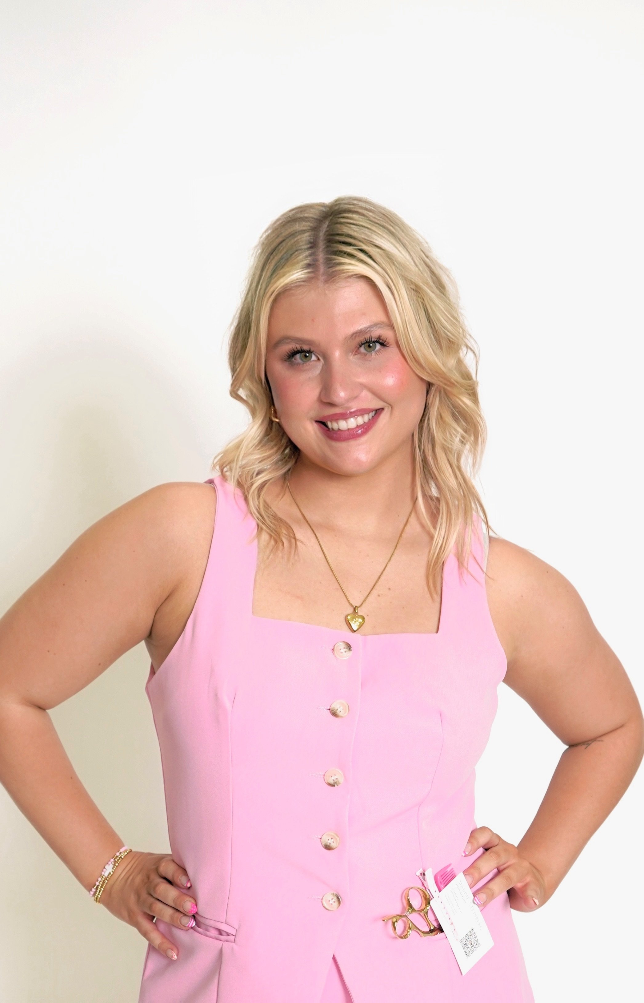 A smiling woman with blonde hair wearing a black sleeveless top with tie straps and white pants standing against a white background.