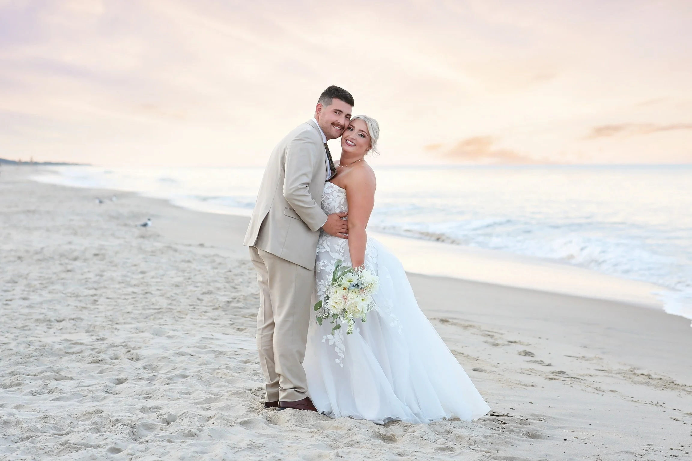 A newlywed couple in wedding attire standing on the beach, smiling, with the ocean and sunset in the background.