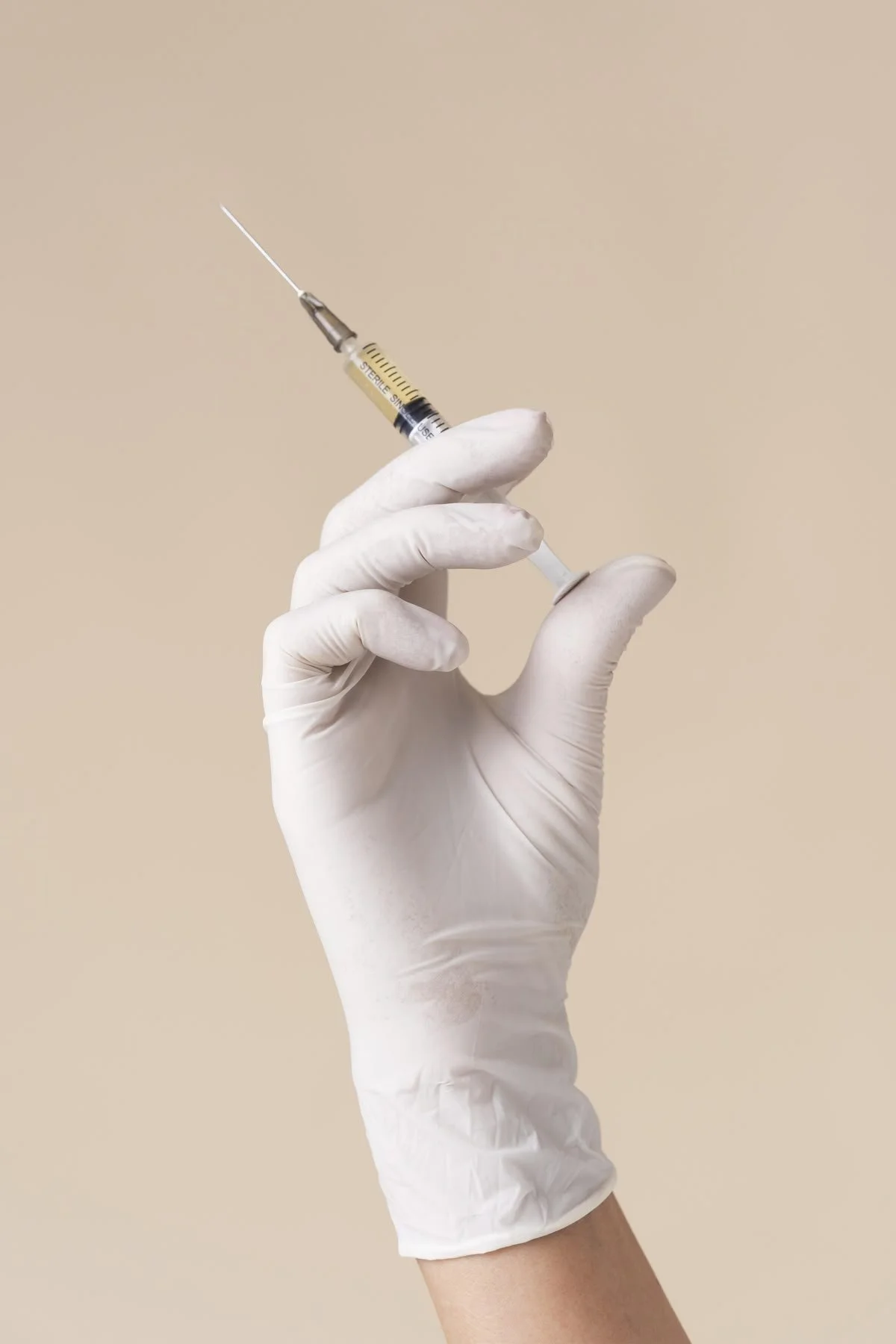 A gloved hand holding a syringe filled with a yellowish liquid against a beige background.