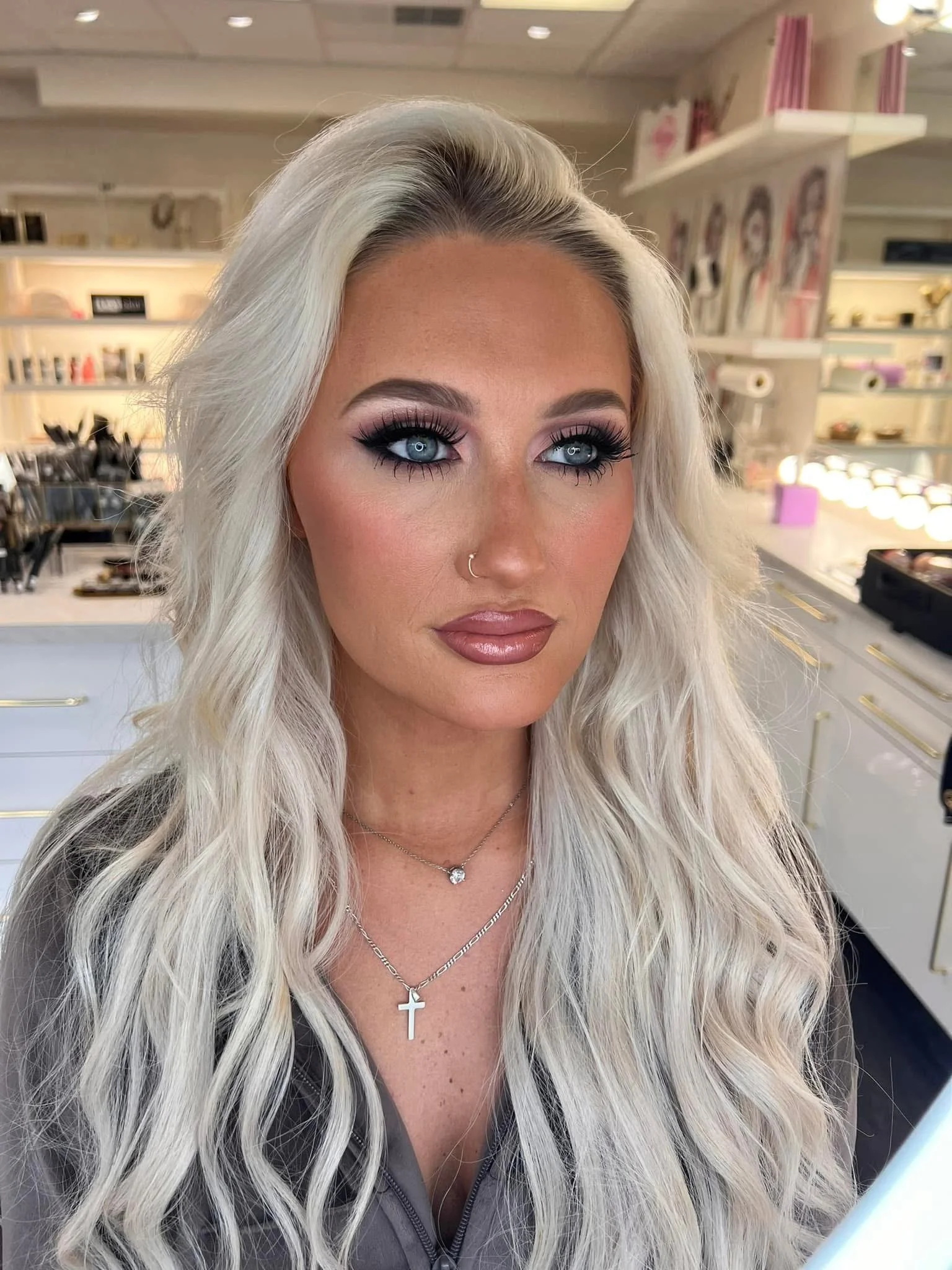 A woman with long, wavy blonde hair and blue eyes, wearing makeup, a nose ring, jewelry, and a gray top, in a makeup studio with shelves of products in the background.