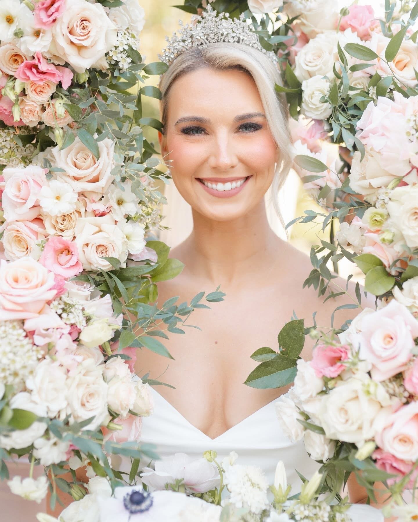 A smiling woman in a wedding dress with a bejeweled headband, framed by a lush arrangement of pink and white roses, greenery, and other flowers.