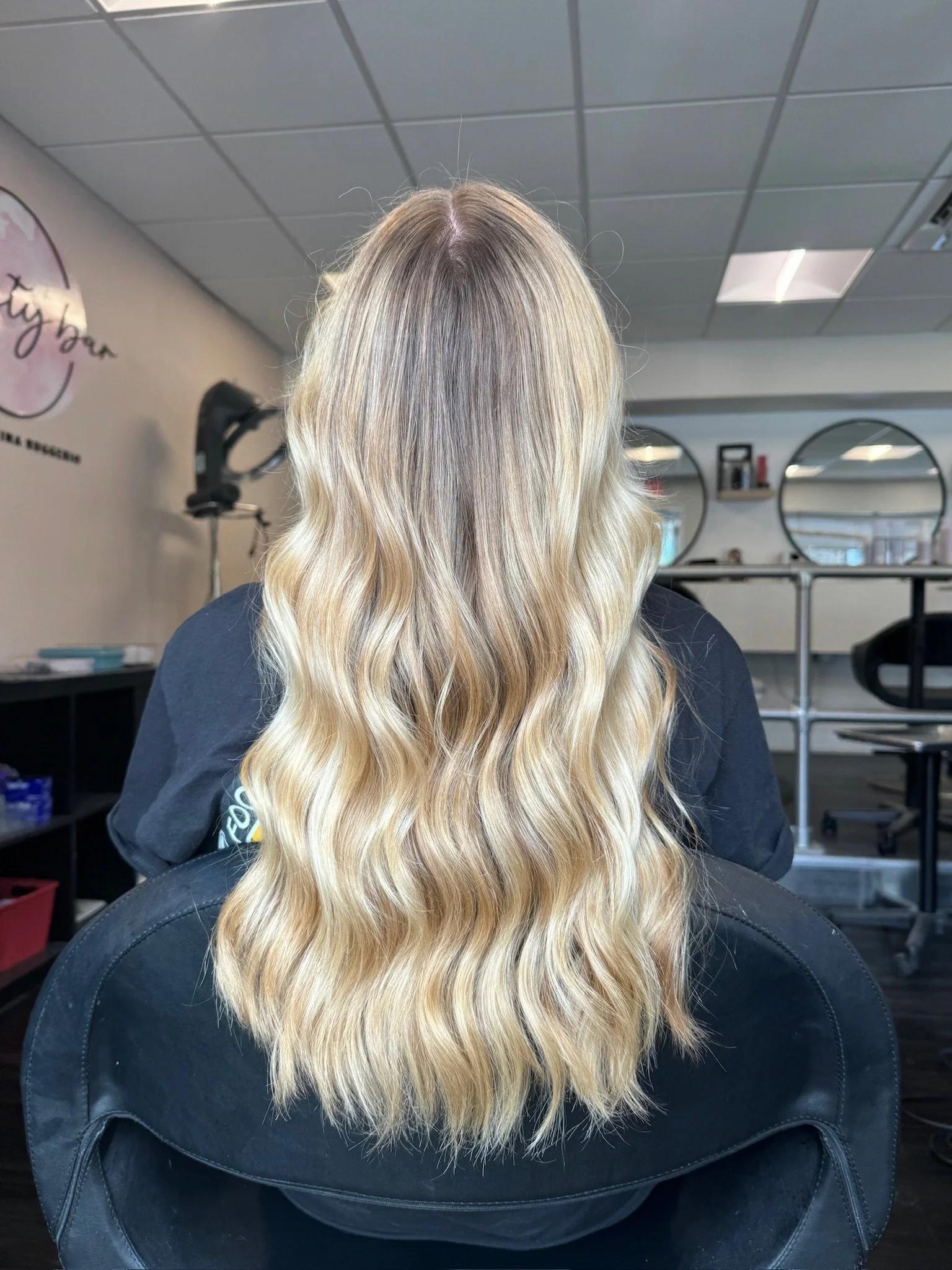 Back view of a woman with long, wavy blonde hair in a salon setting.