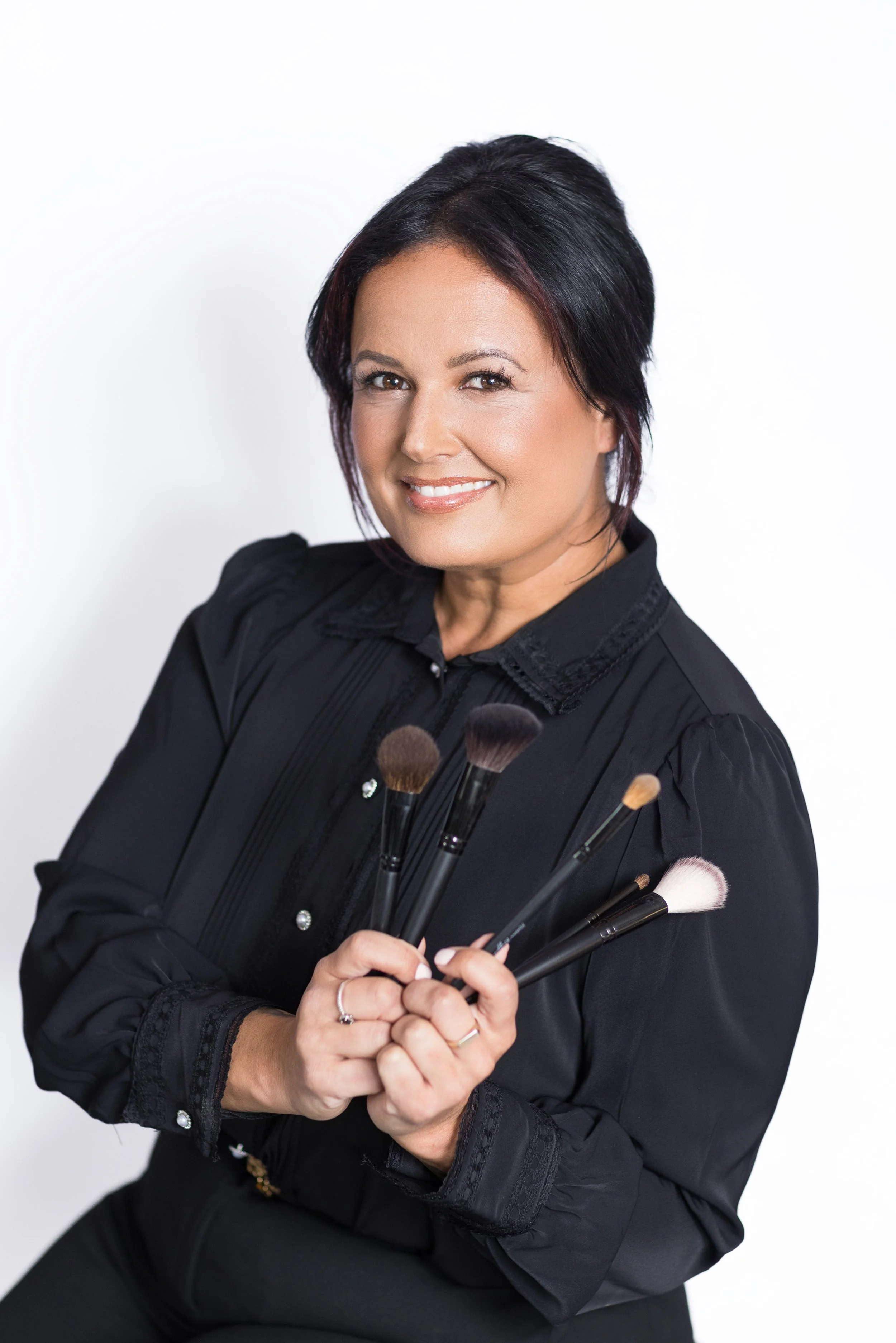 A woman with short black hair holding various makeup brushes, smiling, against a plain white background.