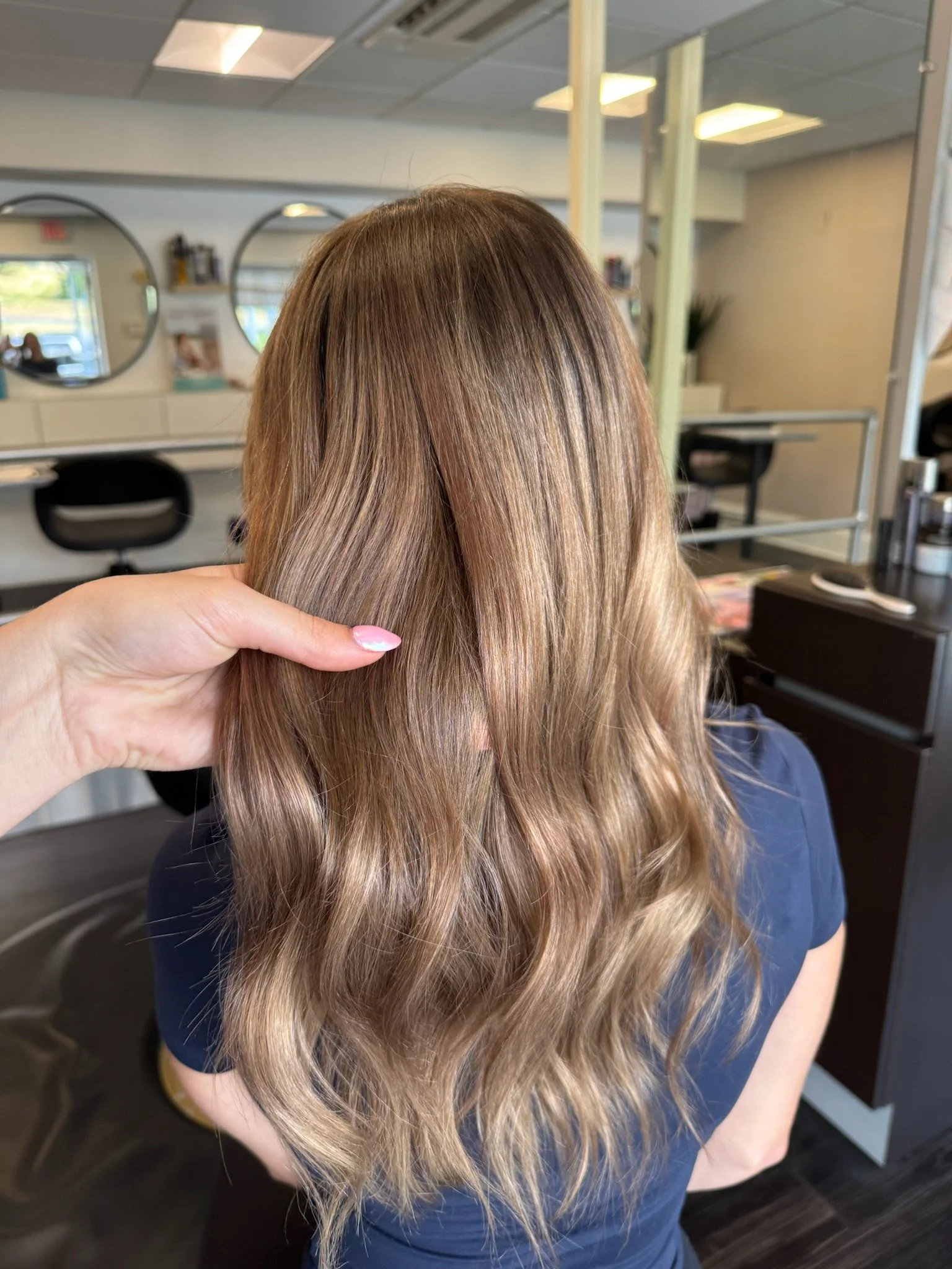 A person with long, wavy, light brown hair in a salon, with an arm holding a section of their hair, and a background of salon mirrors and chairs.